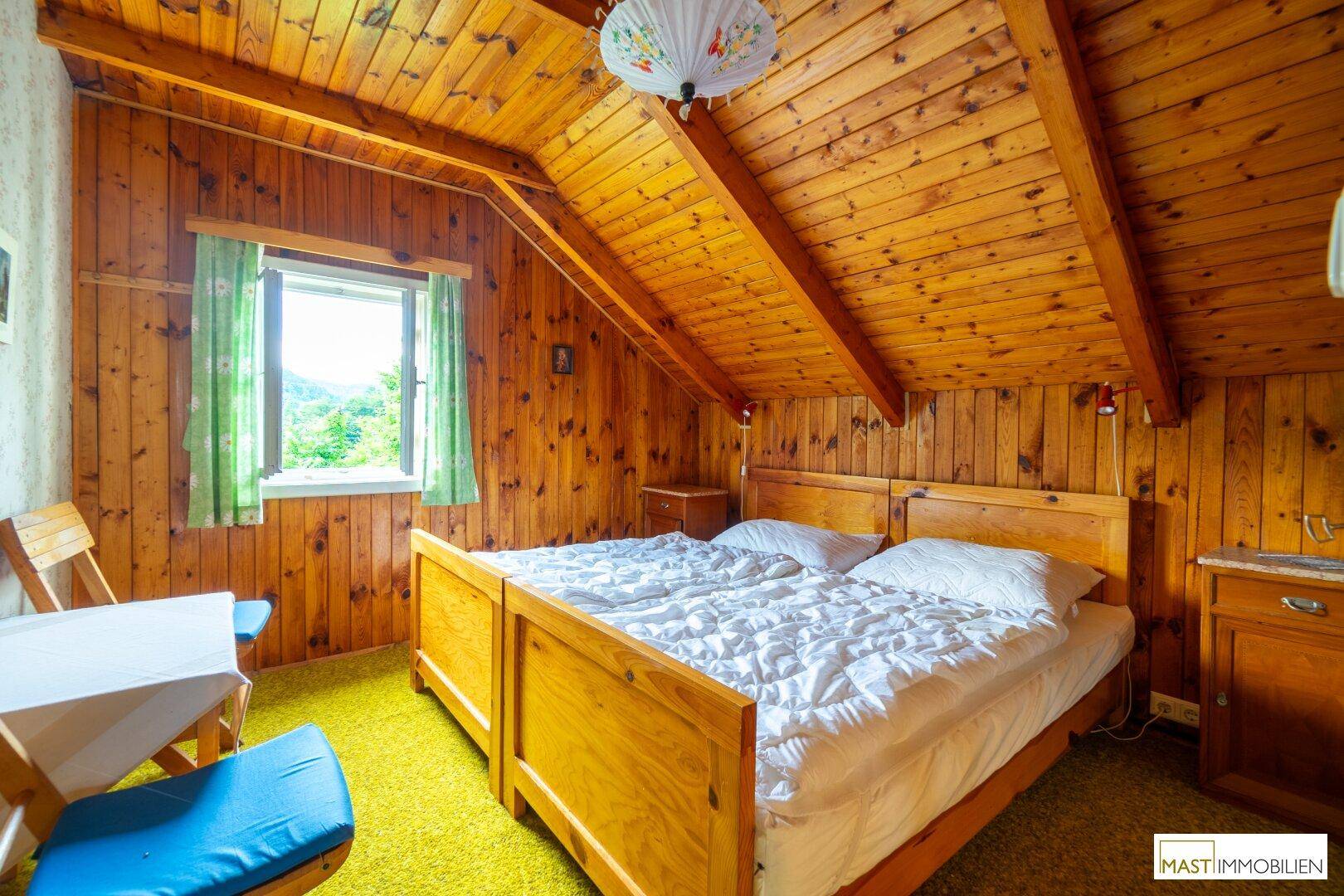 Helles Schlafzimmer mit Holzvertäfelung, schrägen Decken und Blick auf die umliegende Natur.