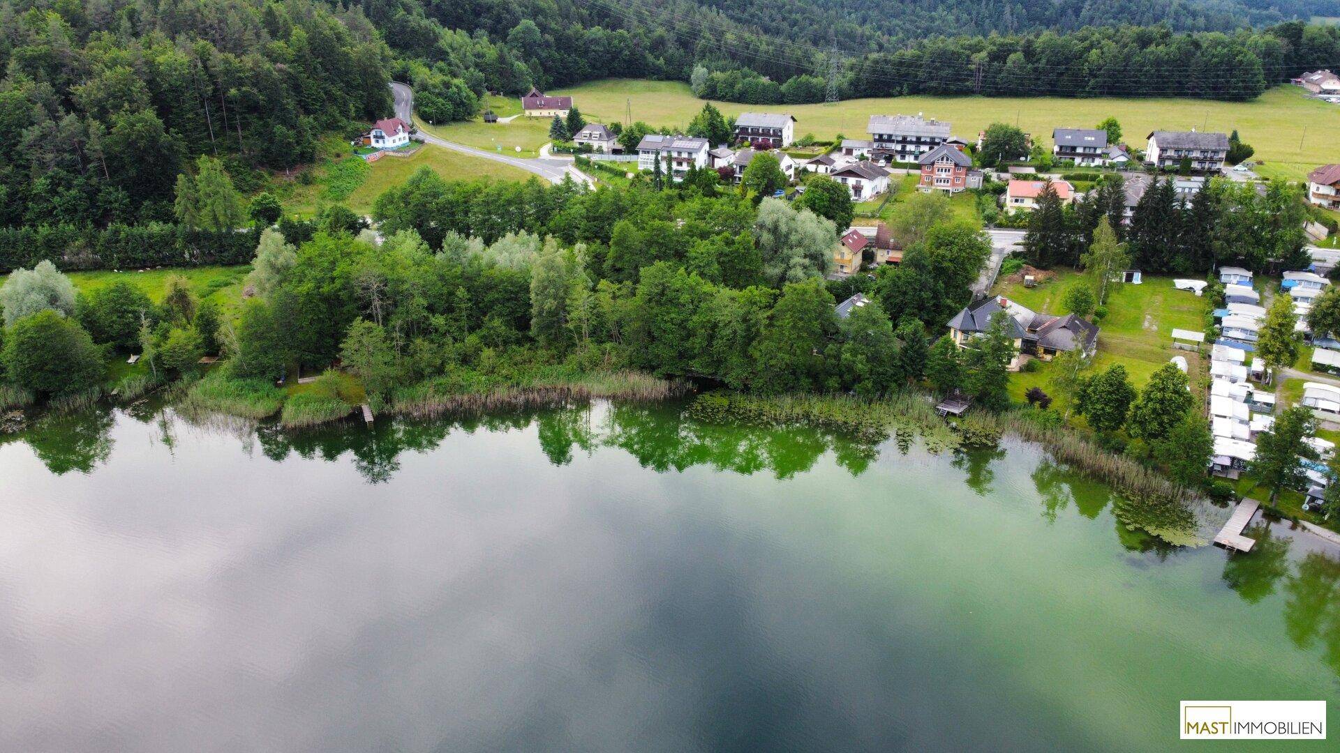 Malerische Luftaufnahme eines Sees mit bewaldeten Ufern und einer kleinen Siedlung im Hintergrund.