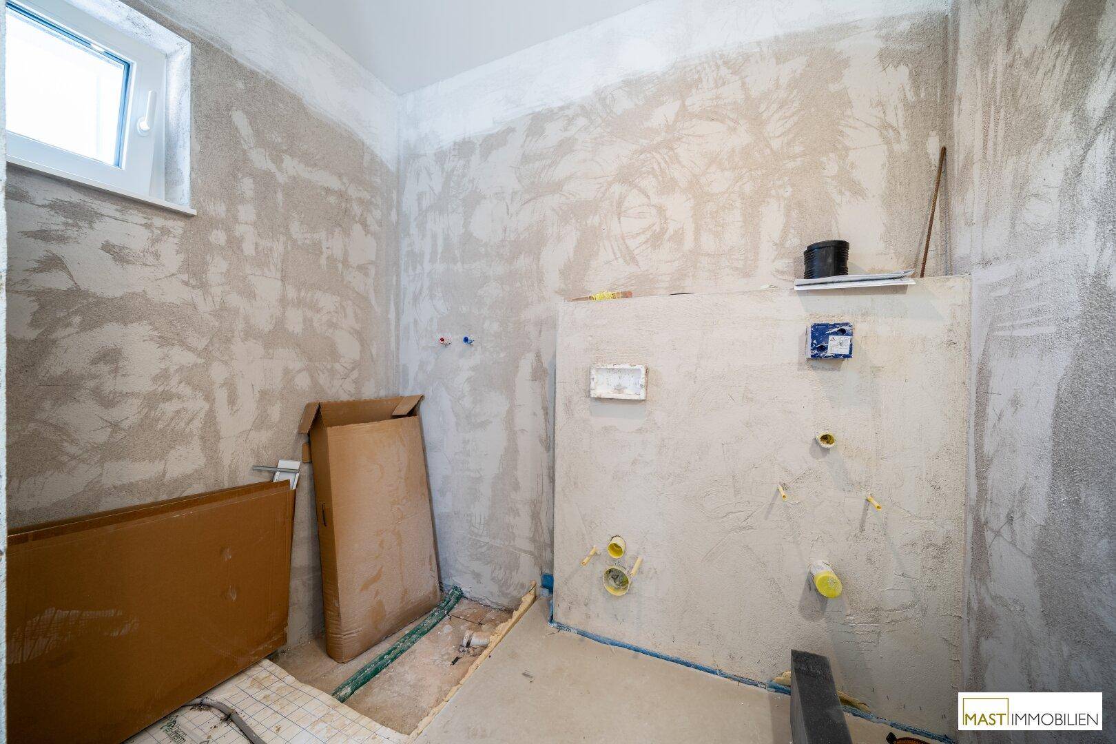 Badezimmer im Rohbauzustand mit sichtbaren Installationen und einem Fenster für Tageslicht.
