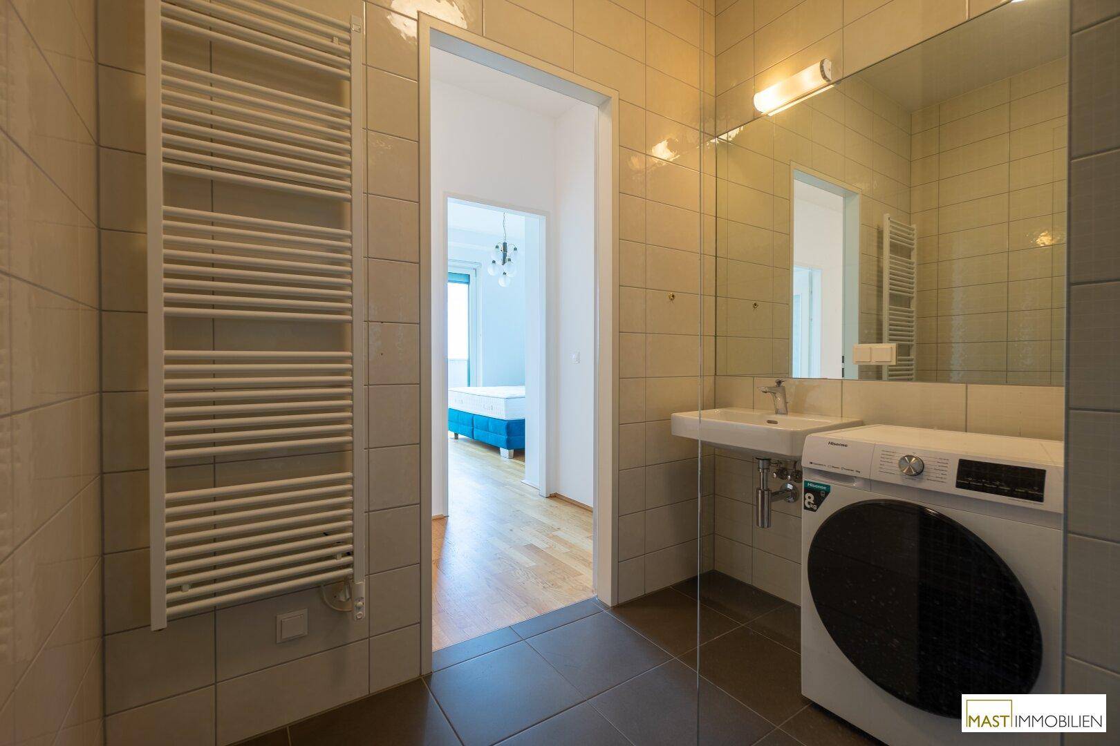 Helles Badezimmer mit Waschmaschine, Handtuchheizkörper und Zugang zu einem Schlafzimmer.