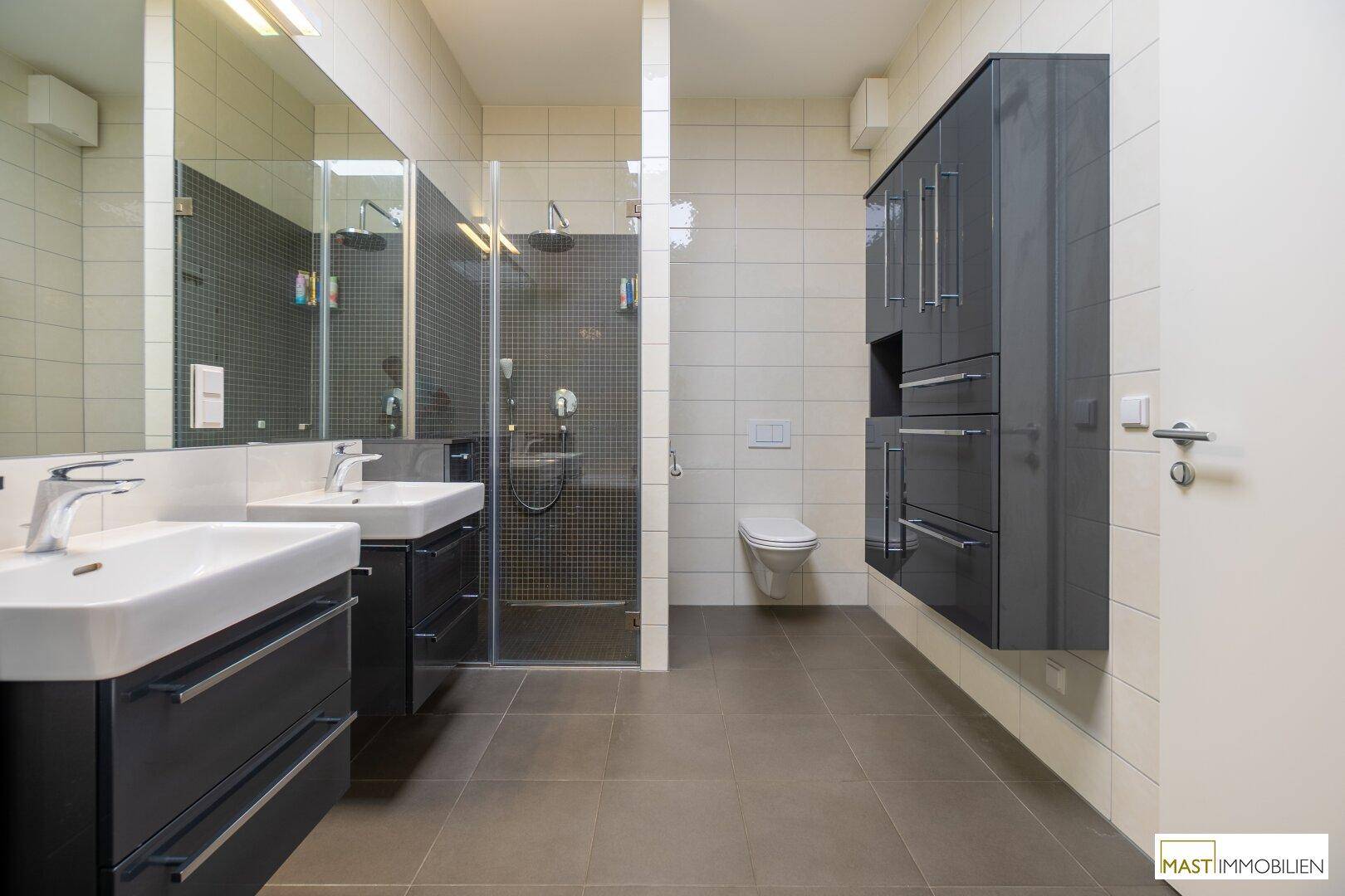 Modernes Badezimmer mit Doppelwaschbecken, großer begehbarer Dusche und Wand-WC, in Grau- und Weißtönen.