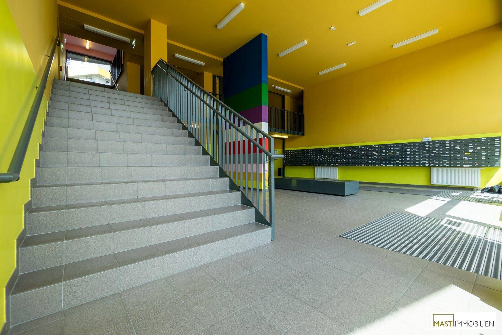 Geräumiger Eingangsbereich mit Treppe, Briefkästen und farbenfroher Gestaltung in Gelb und Blau.