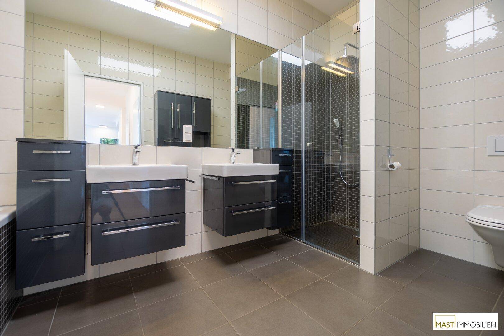 Modernes Badezimmer mit Doppelwaschbecken, begehbarer Dusche und Wand-WC, mit hellen Fliesen.