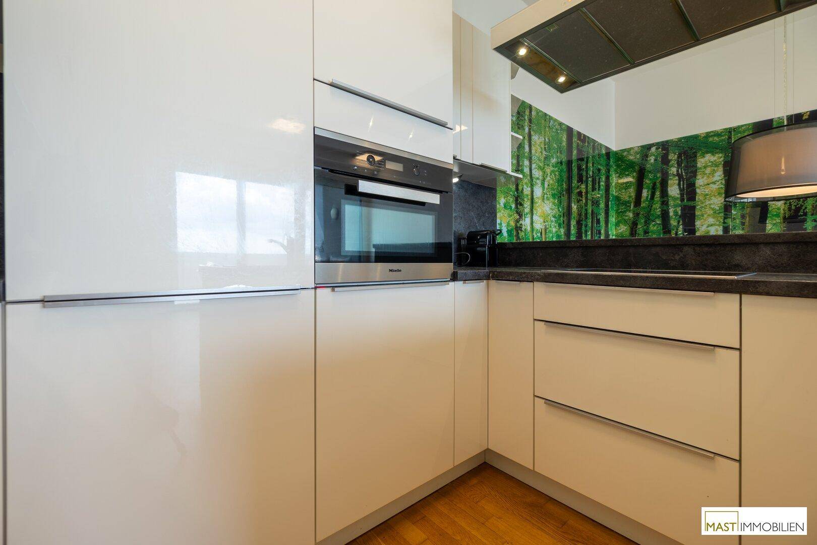 Moderne Einbauküche mit glänzenden weißen Fronten, integriertem Backofen und dunkler Arbeitsplatte.