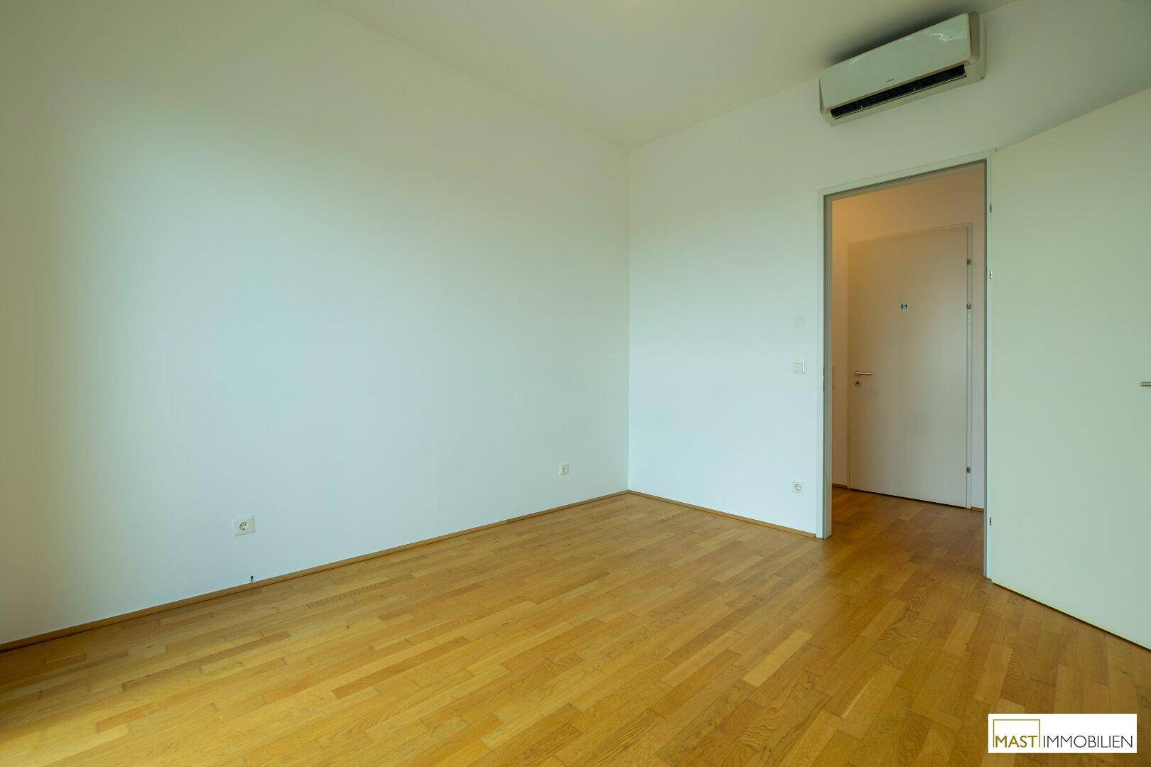 Leeres Zimmer mit Holzboden, weißen Wänden und einer Klimaanlage, bereit zur individuellen Gestaltung.