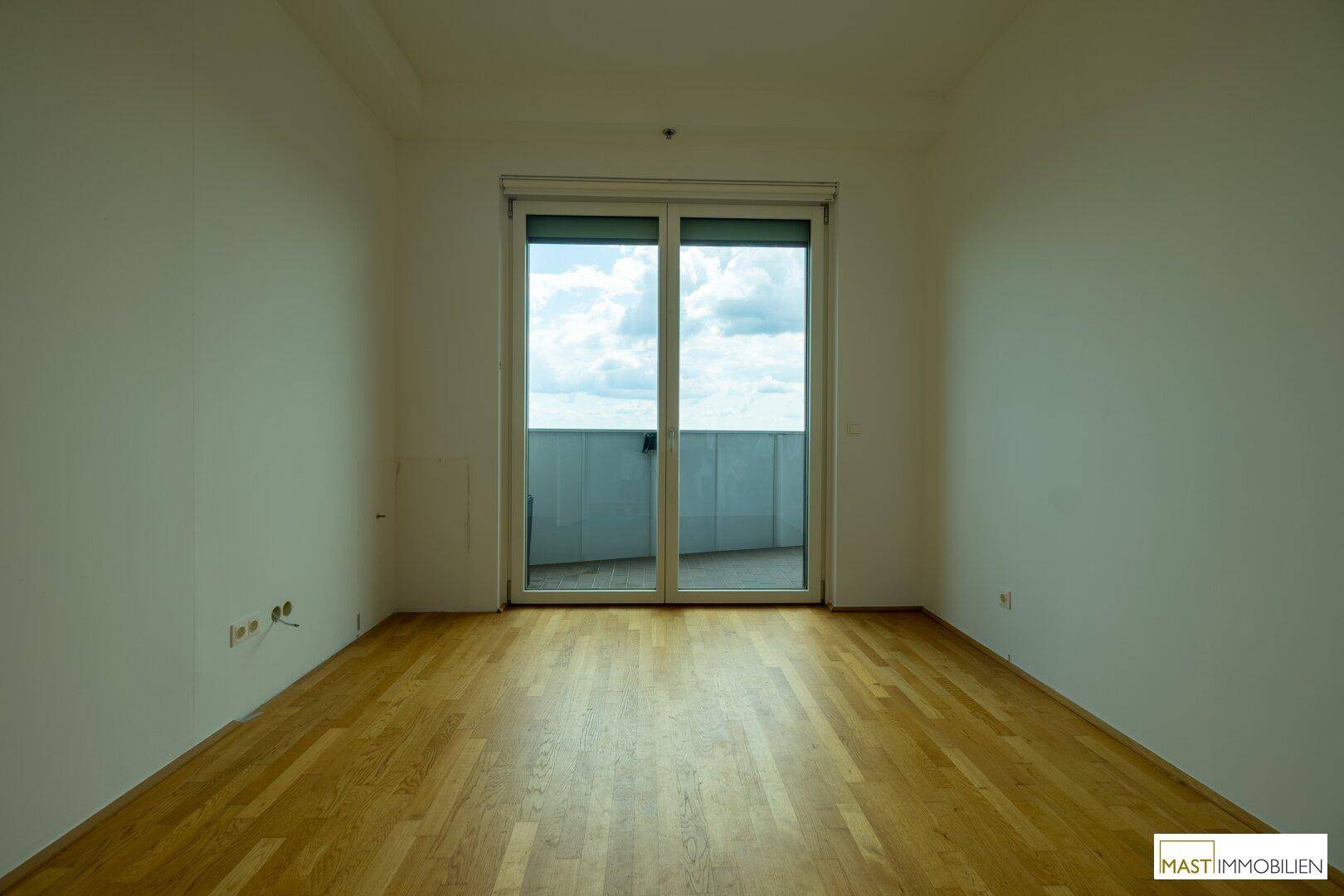 Helles, leeres Zimmer mit Holzboden und großen Schiebetüren, die auf einen Balkon führen.