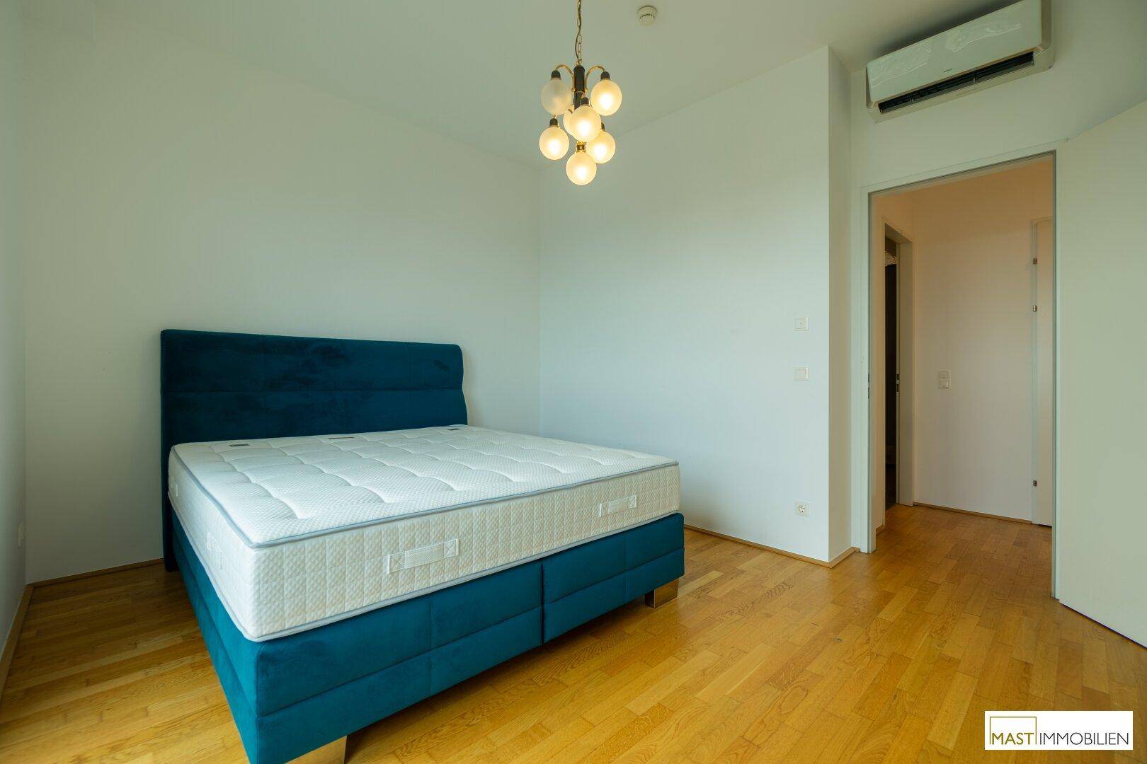 Schlafzimmer mit Holzboden, Klimaanlage und Zugang zu einem weiteren Raum, hell und geräumig.