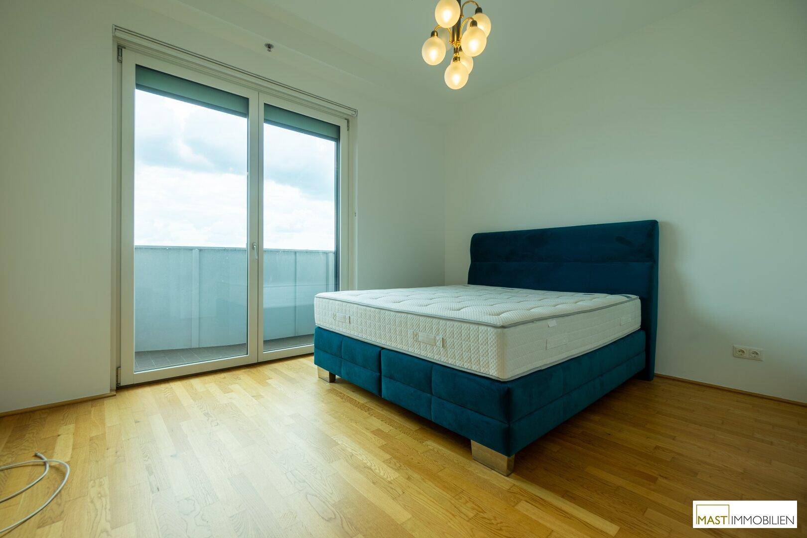 Helles Schlafzimmer mit Holzboden, einem blauen Bett und großen Schiebetüren zum Balkon.