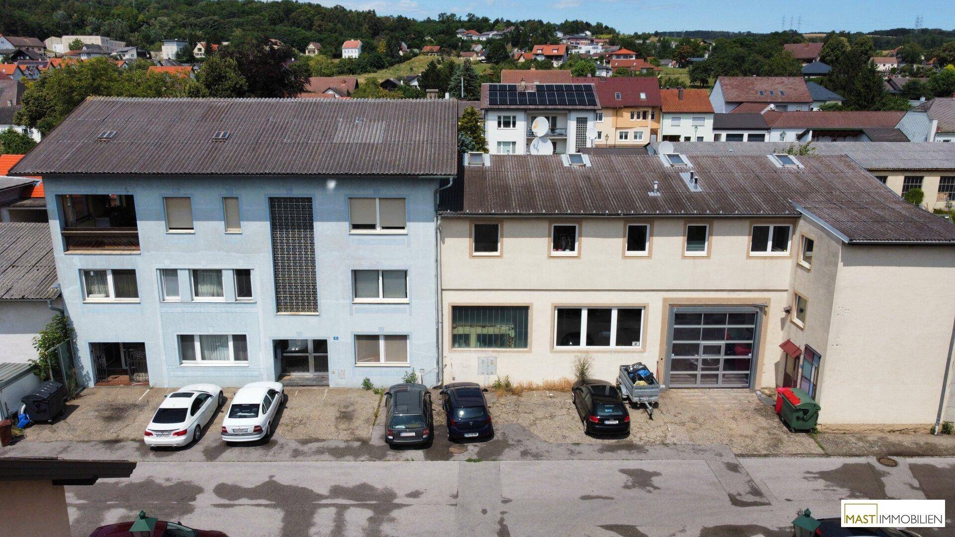 Vorderansicht eines Mehrfamilienhauses mit angrenzendem Gewerbegebäude und Parkplätzen davor.