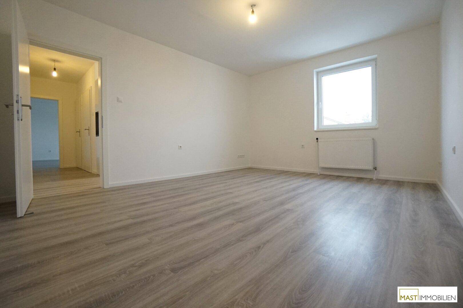 Geräumiger, heller Raum mit Laminatboden, weißen Wänden und einem offenen Durchgang zu einem Flur.