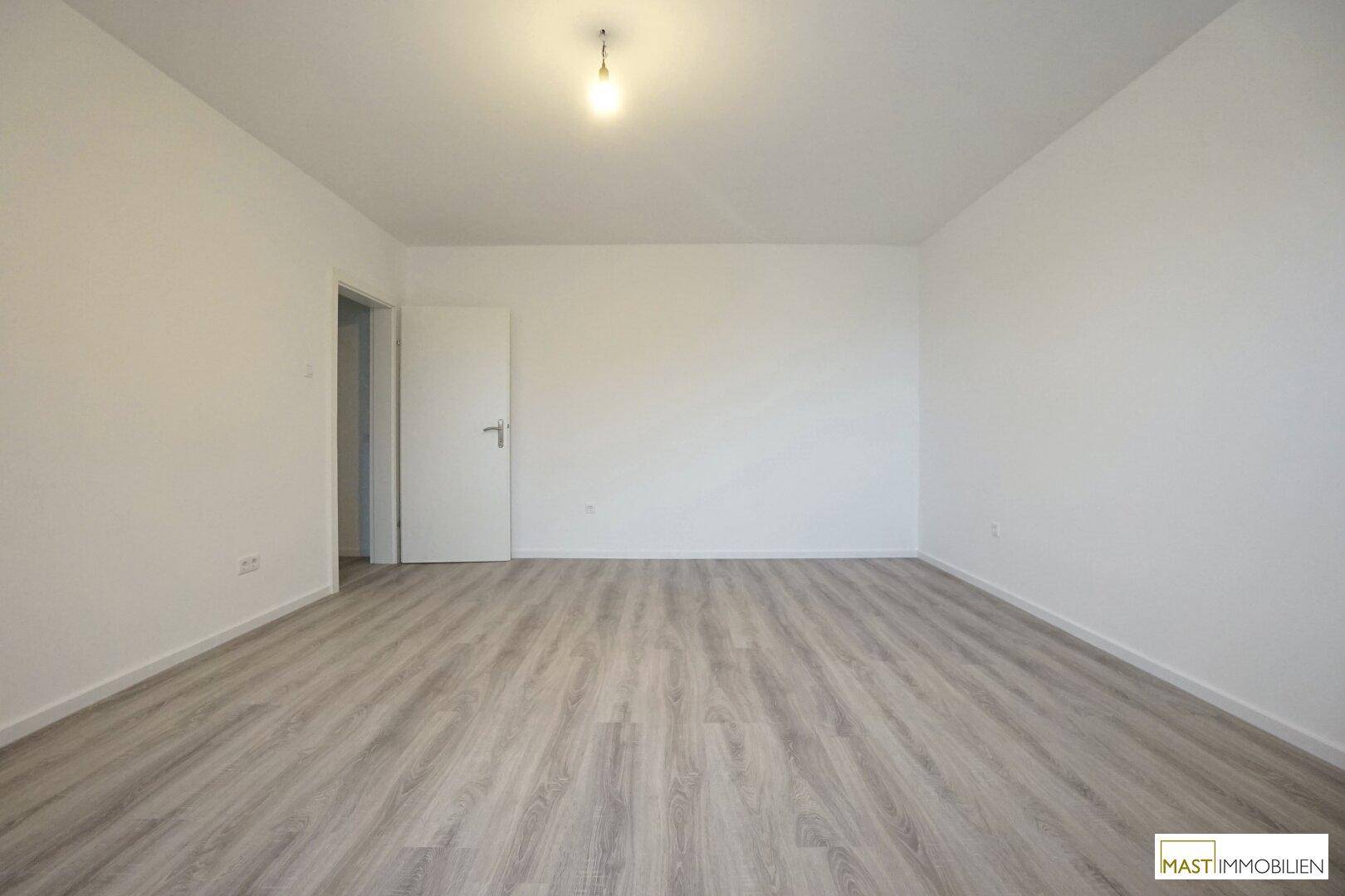 Leerer Raum mit hellem Laminatboden, weißen Wänden und einer geschlossenen Tür, die zu einem anderen Bereich führt.