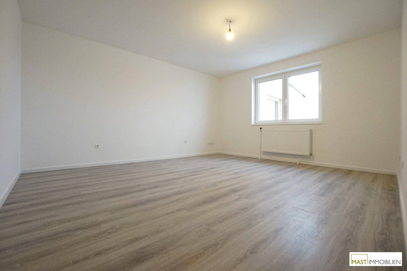 Heller, leerer Raum mit modernem Laminatboden, weißen Wänden und einem Fenster mit Heizkörper.