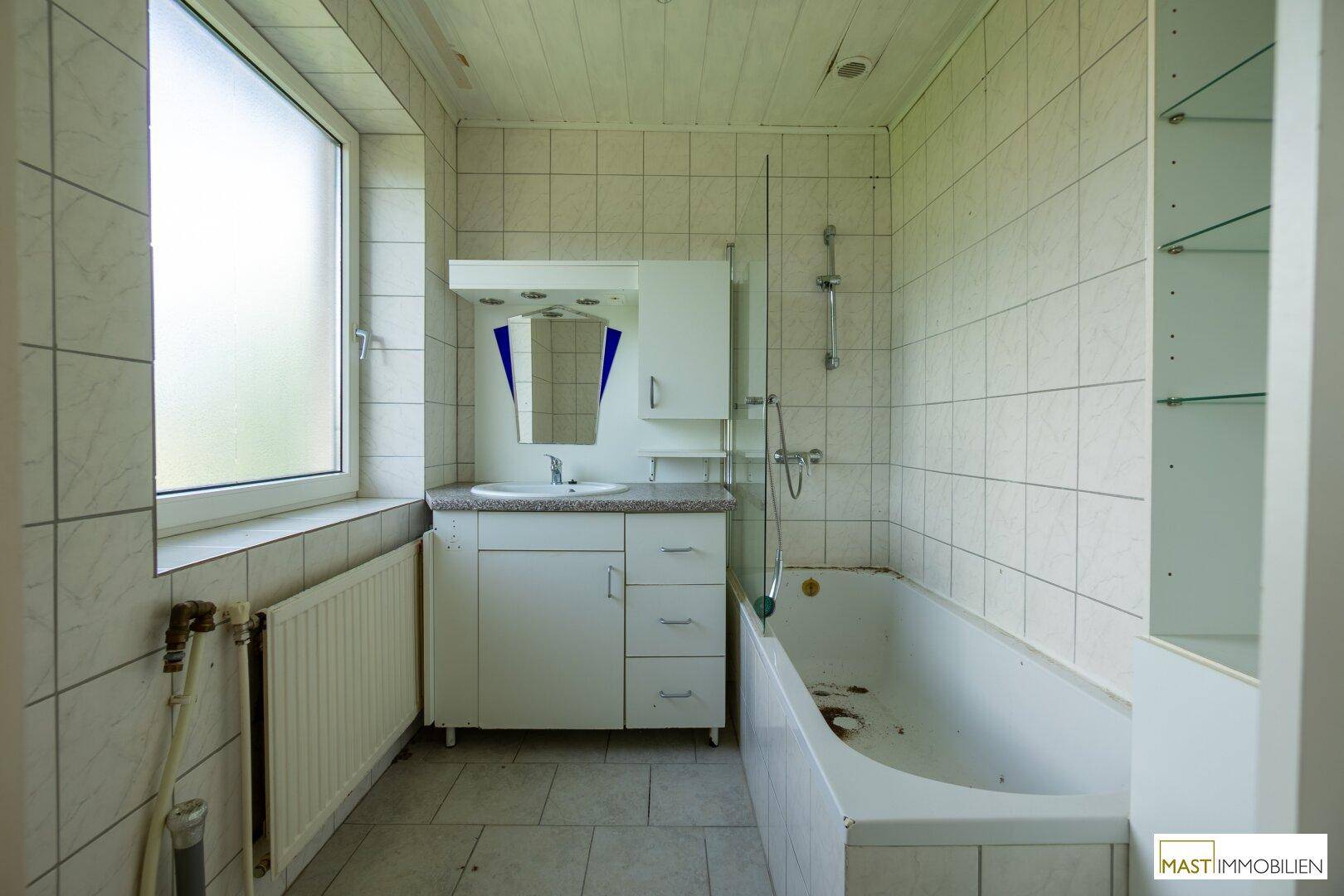 Badezimmer mit Badewanne, Waschbecken, Spiegelschrank und einem Fenster, das Tageslicht hereinlässt.