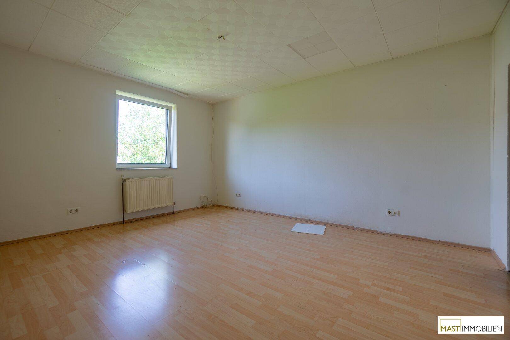 Heller Raum mit Laminatboden, weißen Wänden und einem Fenster, das den Blick ins Grüne freigibt.