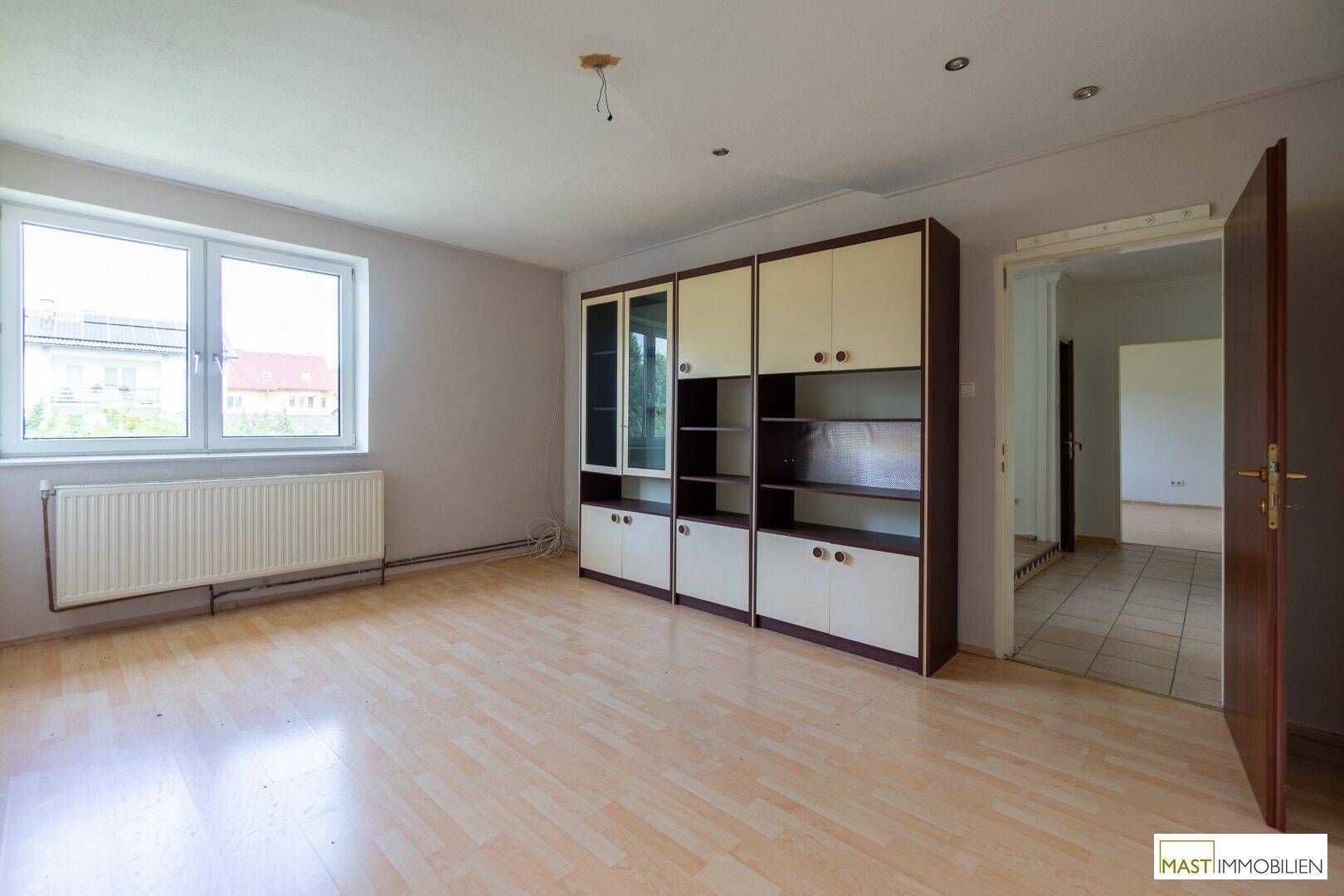 Wohnzimmer mit hellem Laminatboden, großen Fenstern und einem Einbauschrank an der Wand.