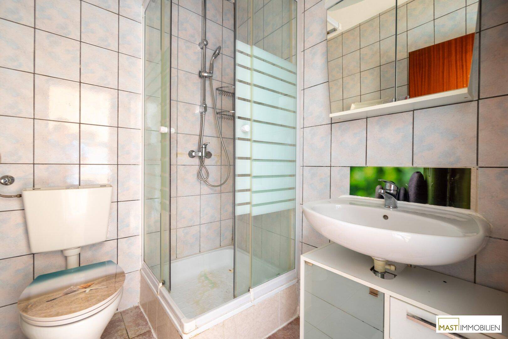 Badezimmer mit Duschkabine, Waschbecken und Toilette, komplett gefliest und mit einem Spiegelschrank.