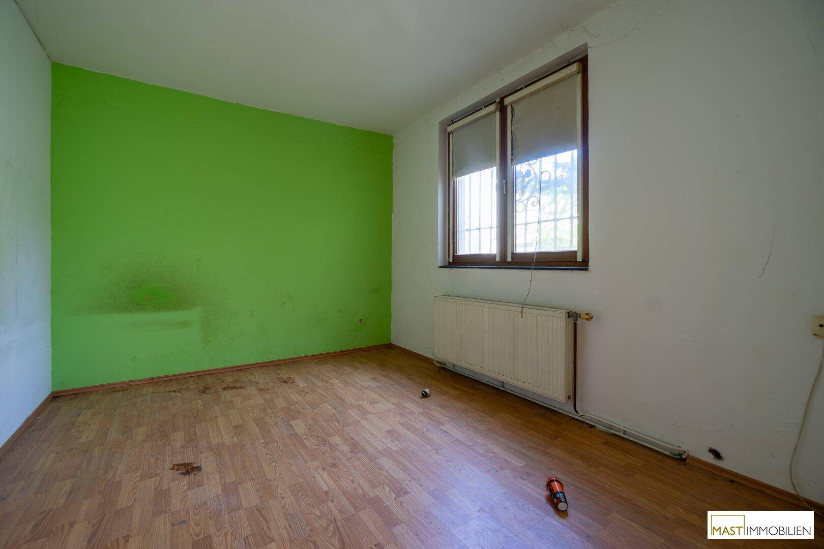 Schlafzimmer mit grüner Akzentwand, Laminatboden und einem Fenster mit Blick ins Grüne.