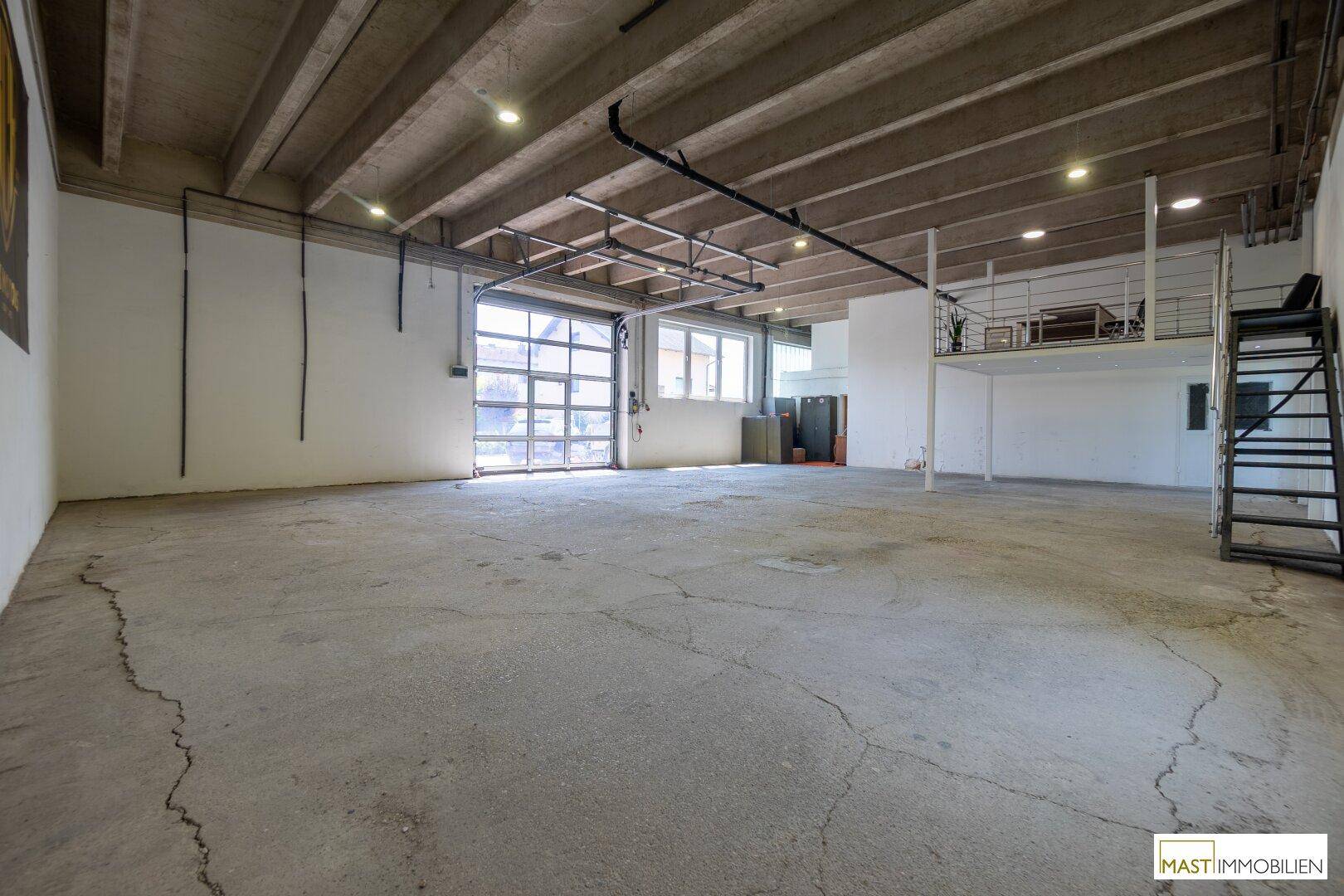 Große Lagerhalle mit Betonboden, hohen Decken und einem breiten Rolltor für Fahrzeuge.