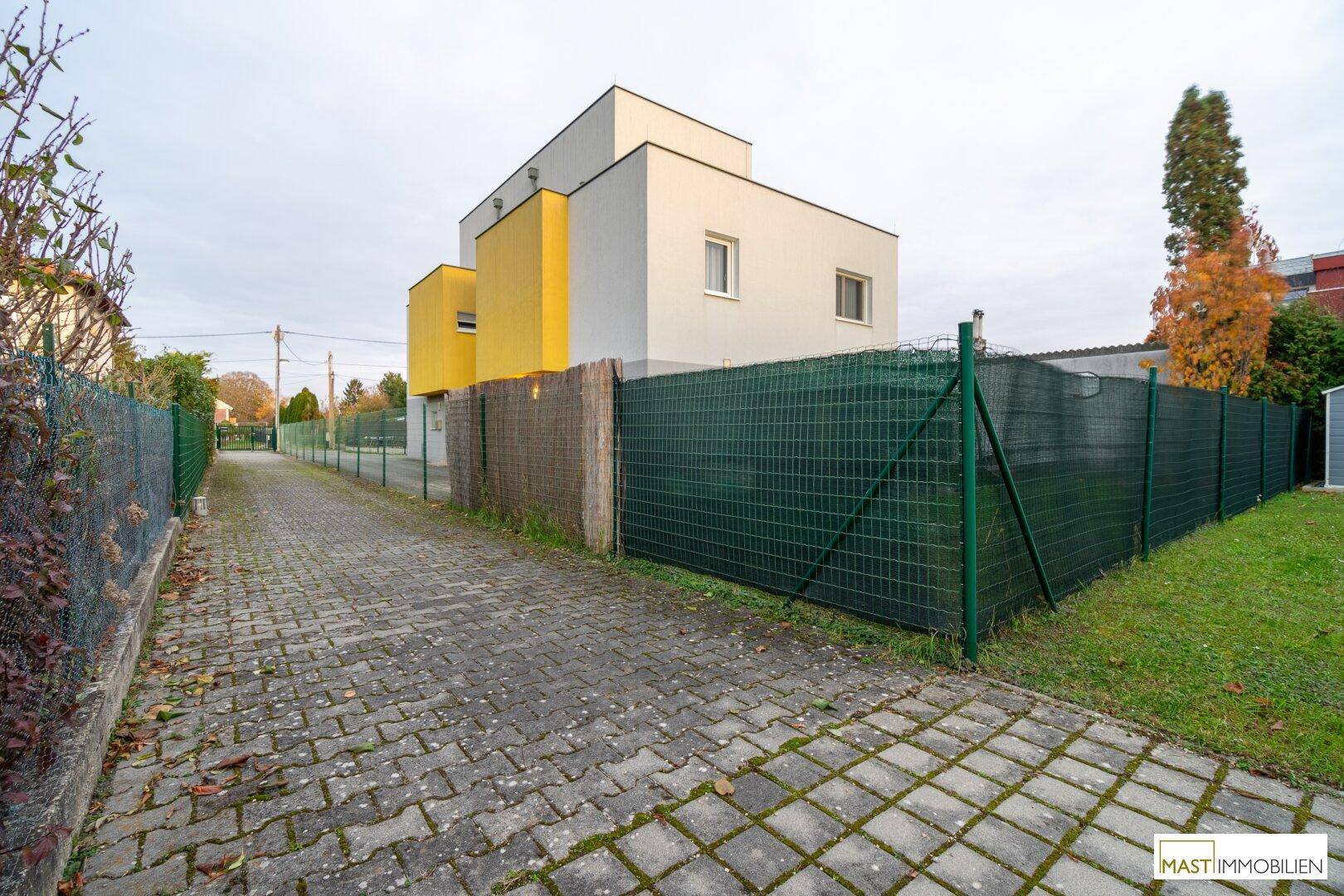 Zufahrt zum Haus mit gepflastertem Weg und grünem Zaun.