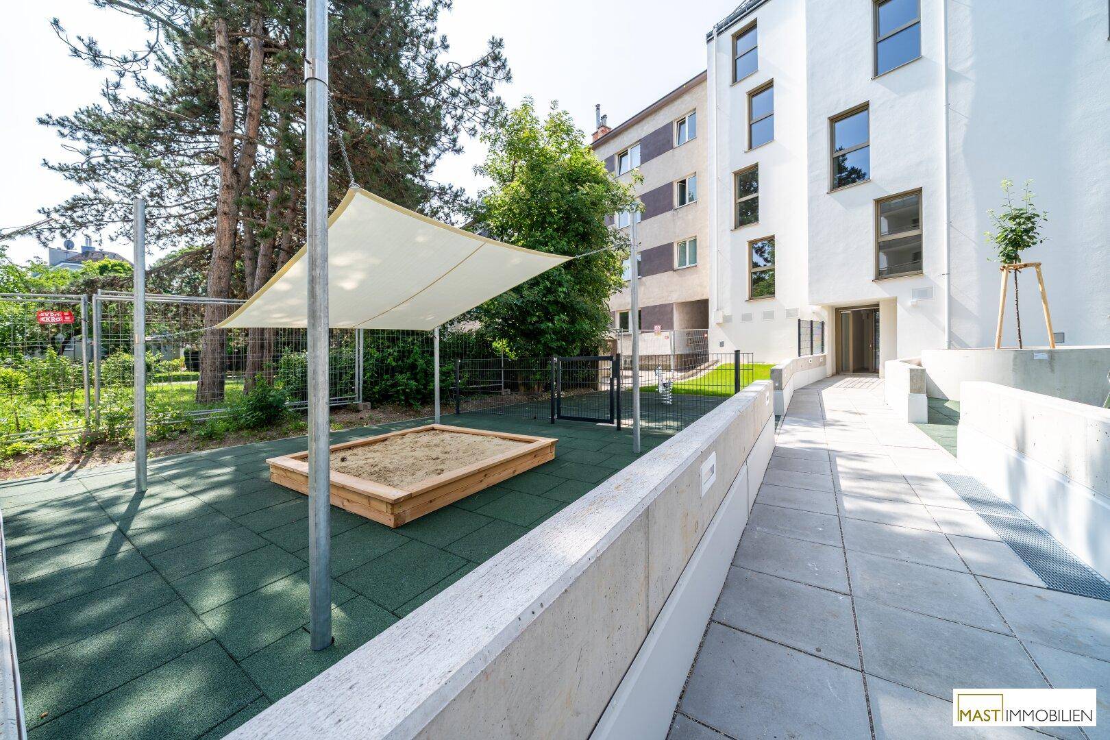 Gemeinschaftsgarten mit einem Sandkasten und Sonnensegel, umgeben von Bäumen und Wohngebäuden.