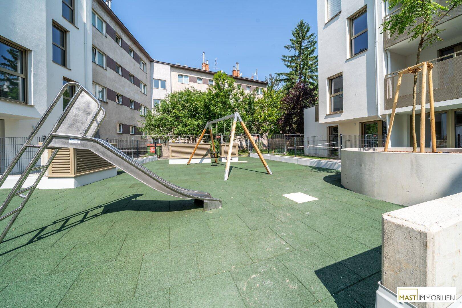 Kinderspielplatz mit Rutsche und Schaukeln auf grünem Fallschutzboden neben einem modernen Wohnhaus.
