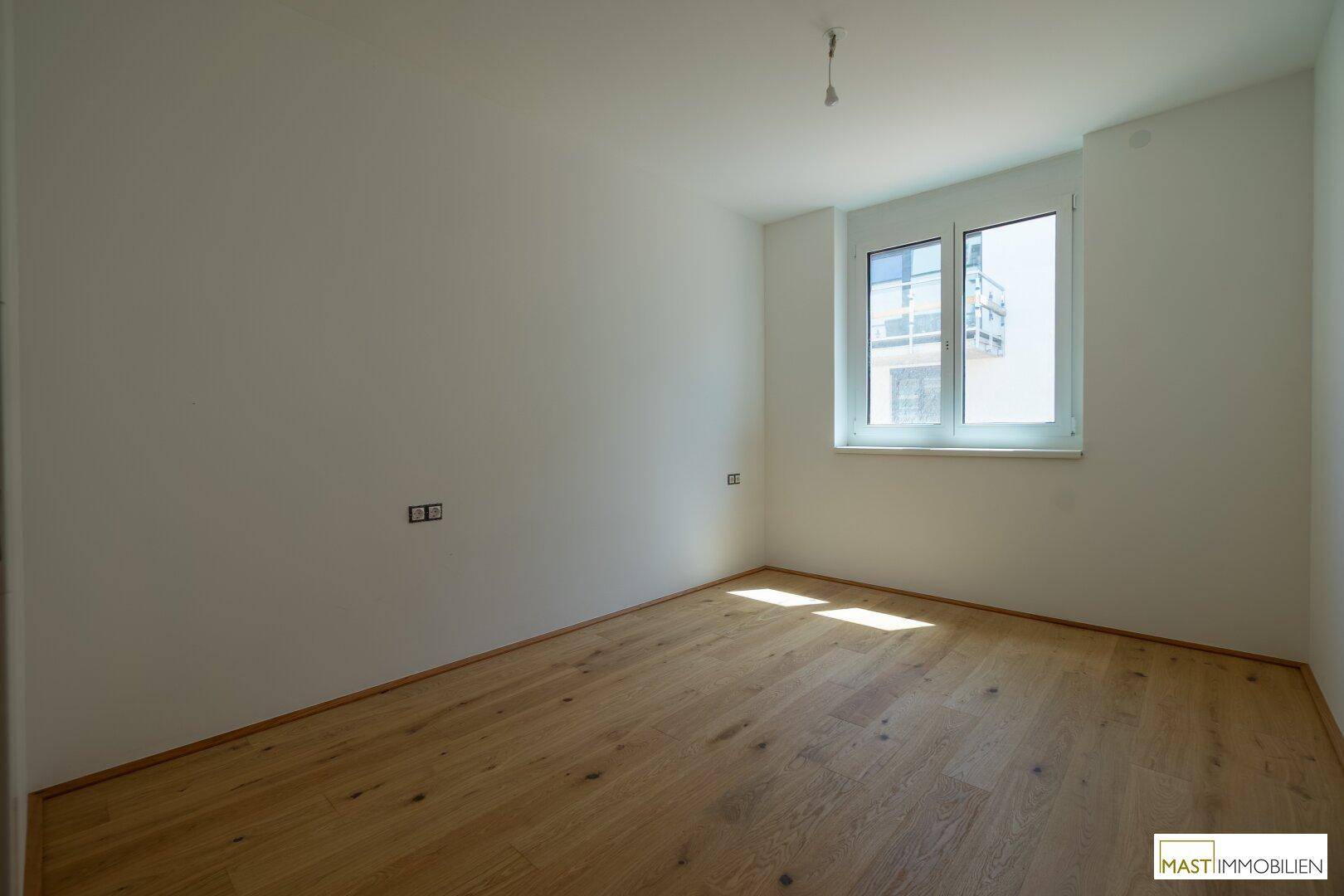 Leeres Schlafzimmer mit hellem Parkettboden und einem Fenster, das viel Licht hereinlässt.