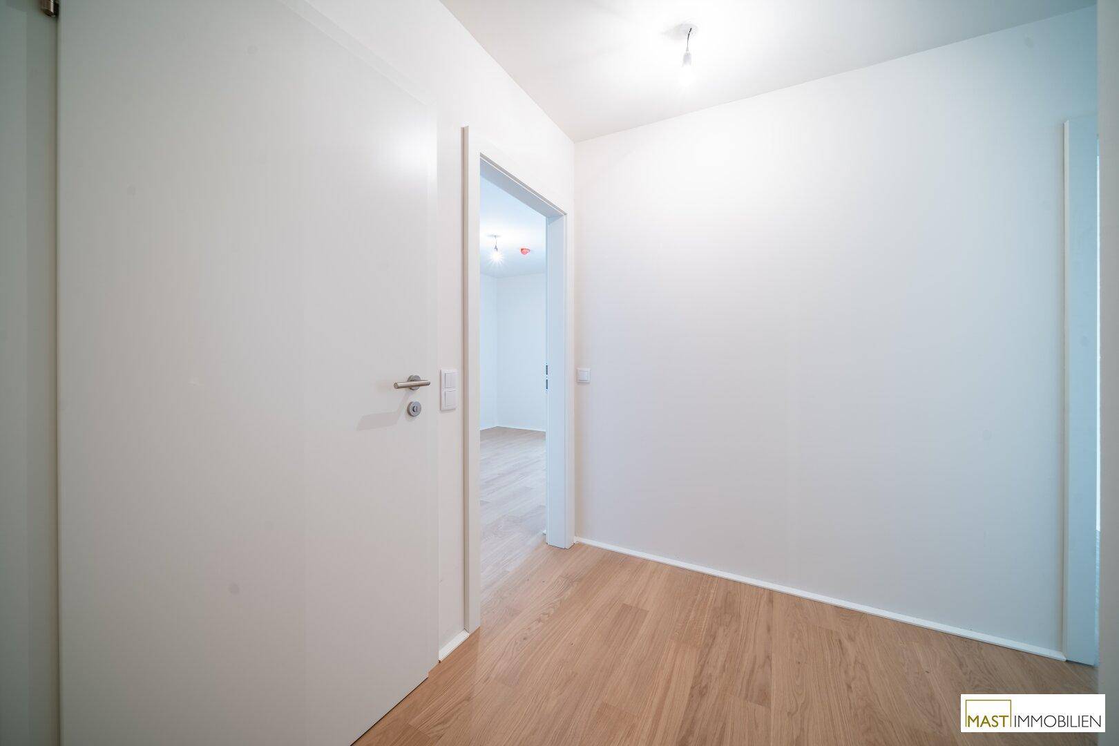 Heller Flur mit Holzboden und weißen Wänden, der einen Blick in einen angrenzenden Raum bietet.