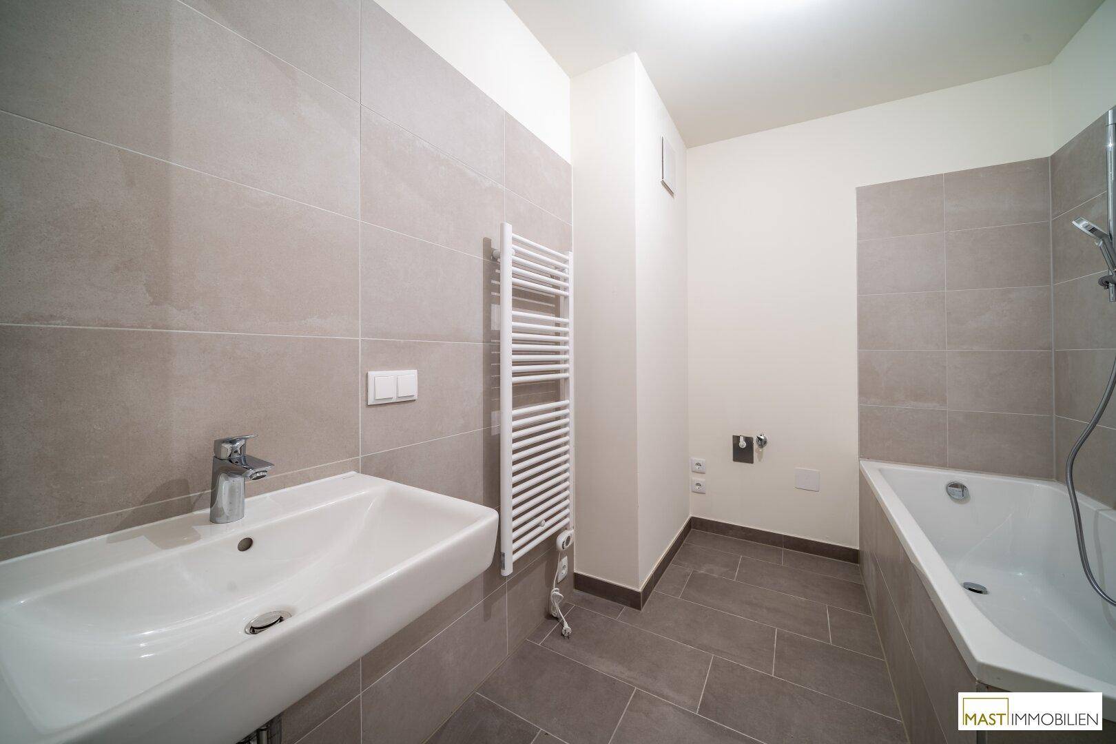 Modernes Badezimmer mit grauen Fliesen, Badewanne, Waschbecken und beheiztem Handtuchhalter.