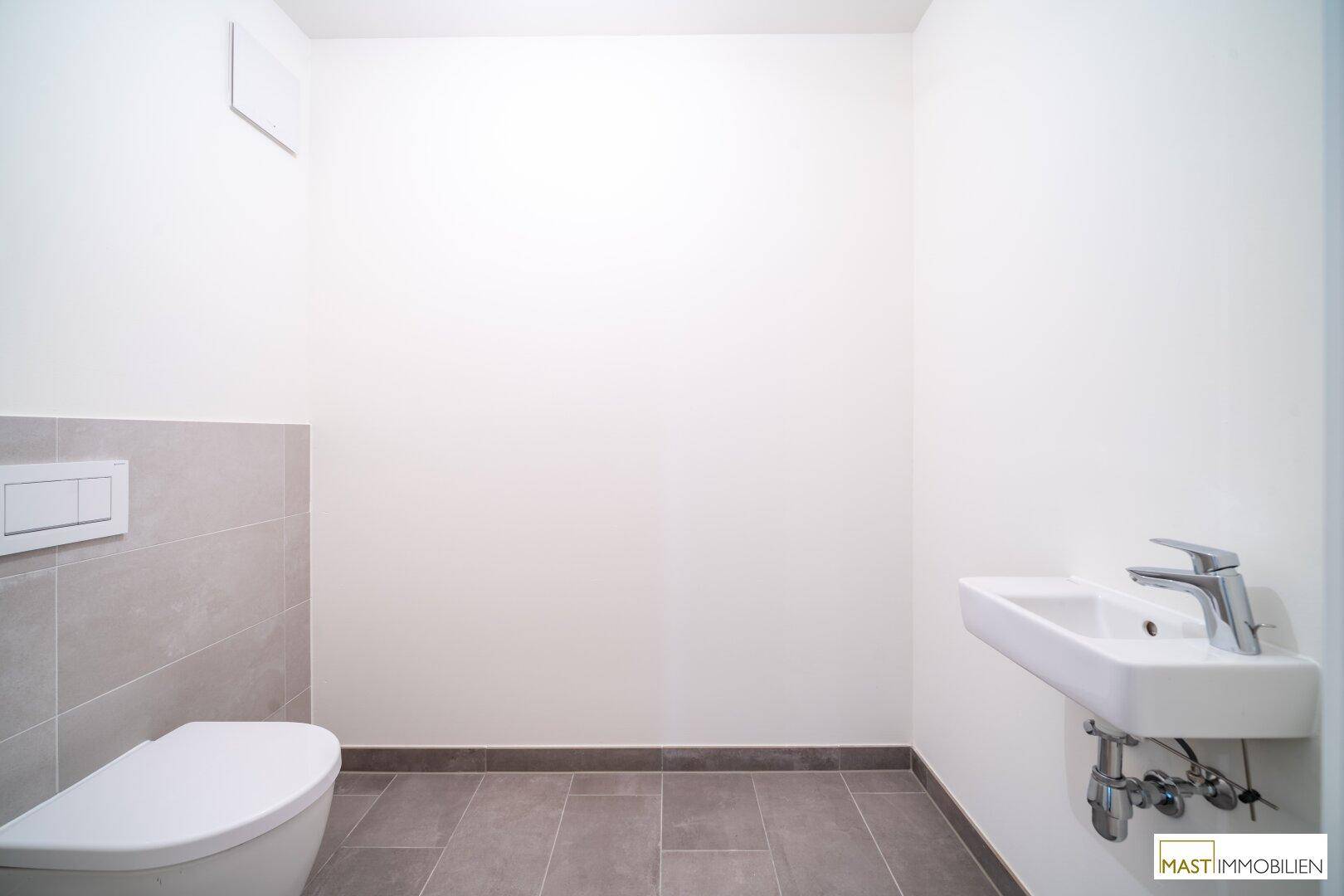 Separates WC mit modernen grauen Fliesen, wandhängender Toilette und kleinem Handwaschbecken.