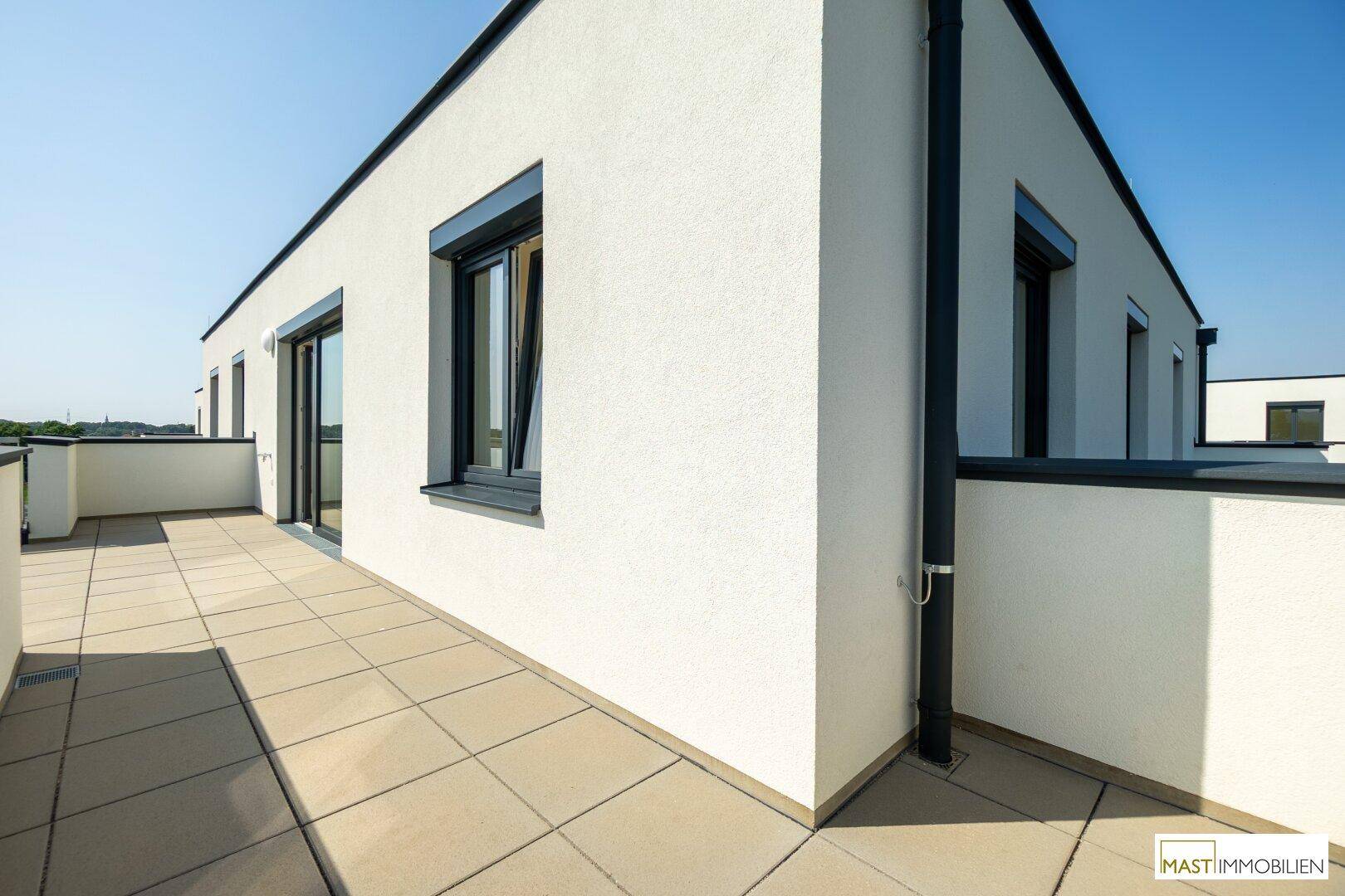 Moderne Gebäudefassade mit weißem Putz und einer großzügigen Dachterrasse unter blauem Himmel.