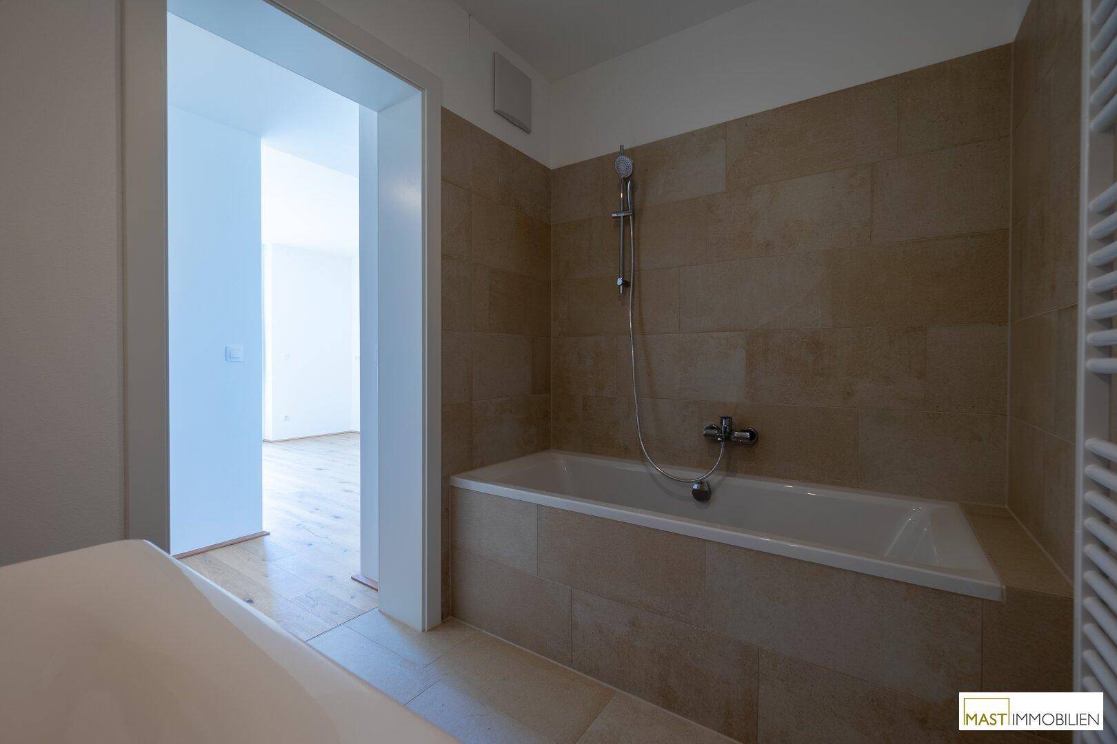 Badezimmer mit Badewanne und beigefarbenen Fliesen, mit Blick in einen angrenzenden Raum.
