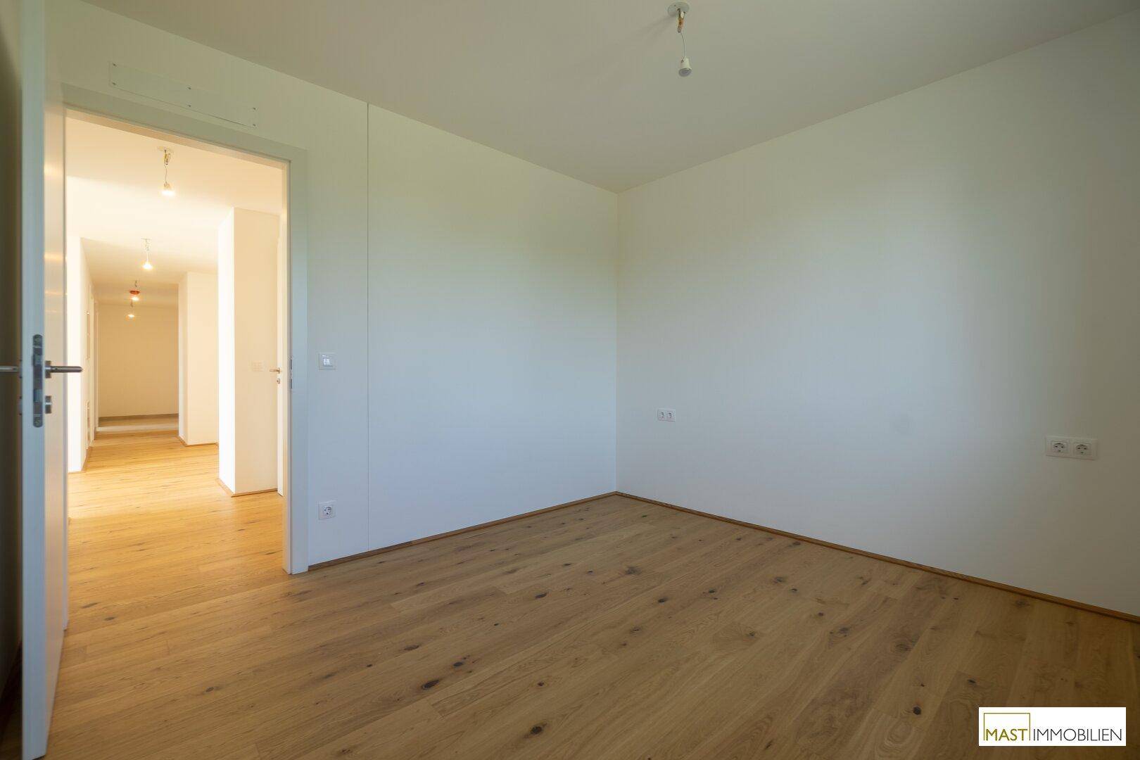 Leeres Zimmer mit Holzboden und Zugang zu einem langen, hellen Flur der Wohnung.