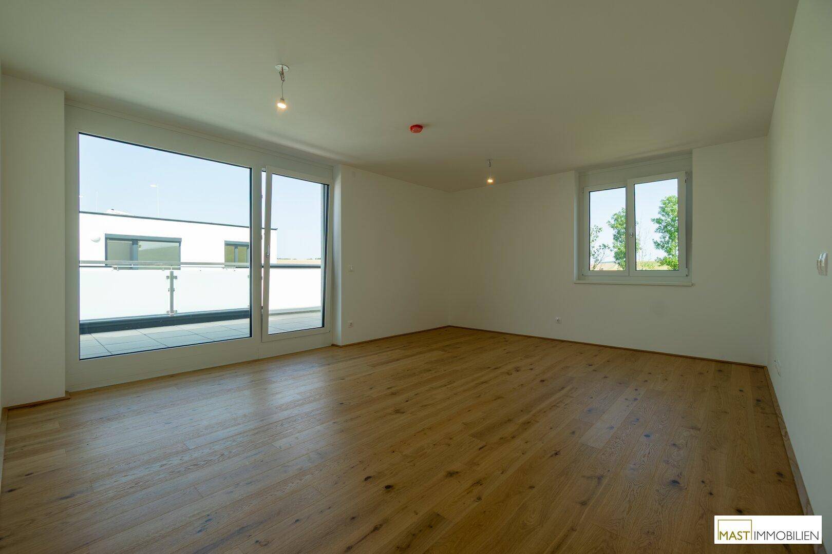 Heller, leerer Raum mit Holzboden und großen Fenstern, die viel Tageslicht hereinlassen.