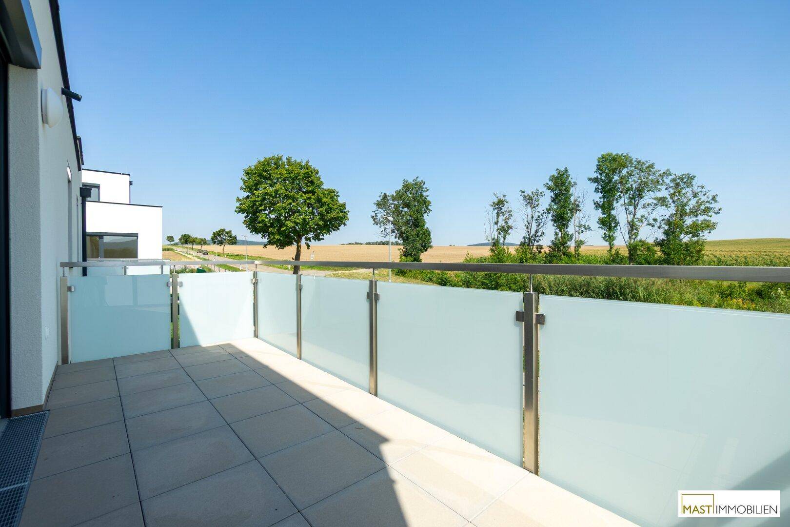 Großer Balkon mit Fliesen und Glasgeländer, umgeben von Feldern und Bäumen unter blauem Himmel.