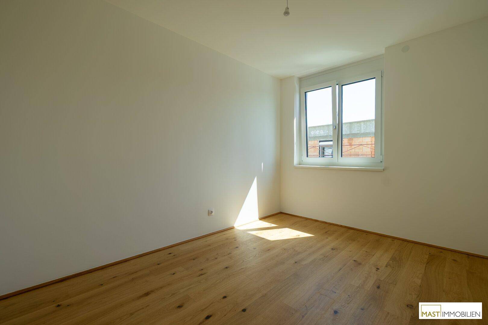 Leeres Schlafzimmer mit hellem Holzparkett und großem Fenster, das viel Sonnenlicht hereinlässt.