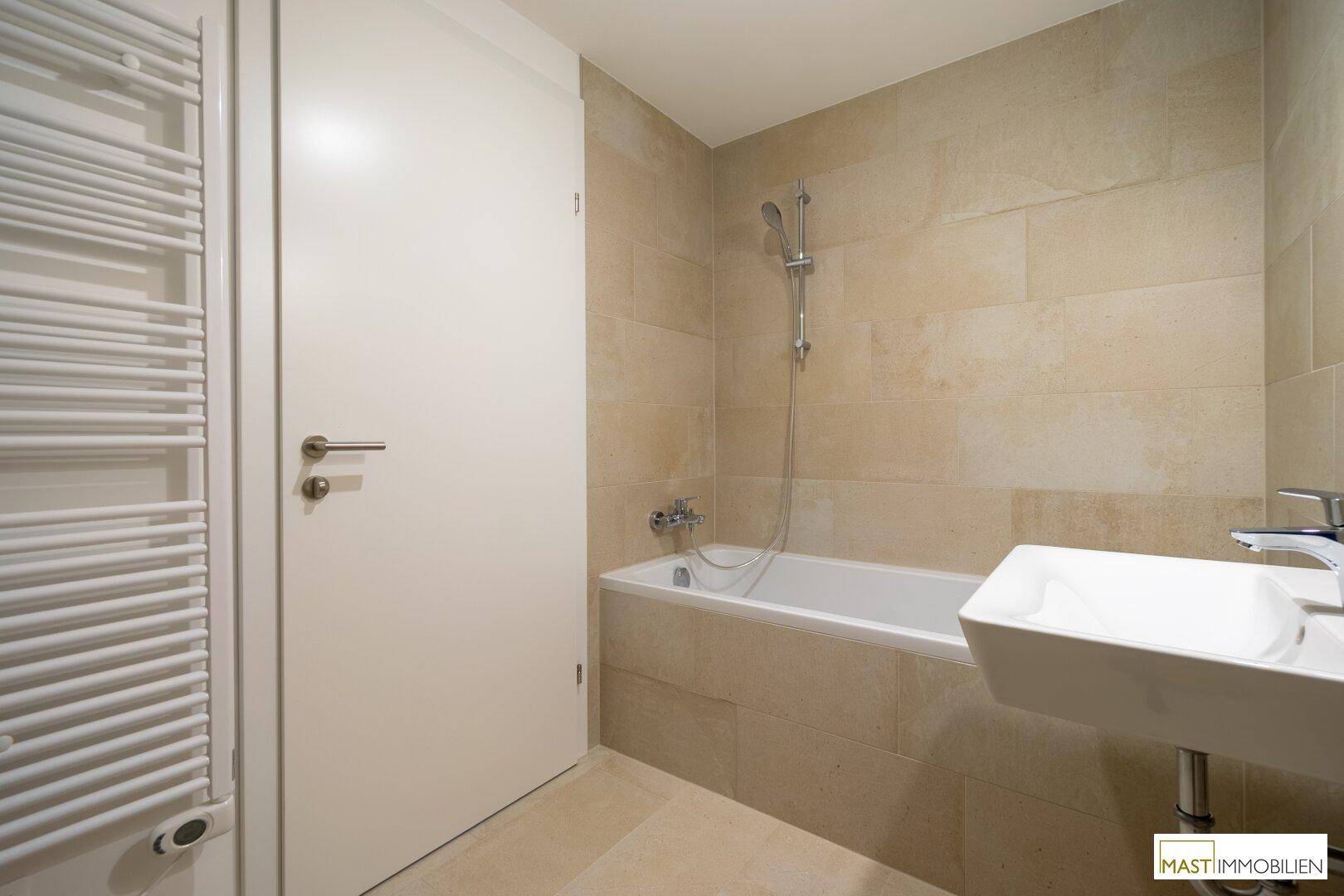 Modernes Badezimmer mit beigefarbenen Fliesen, Badewanne und beheiztem Handtuchhalter.