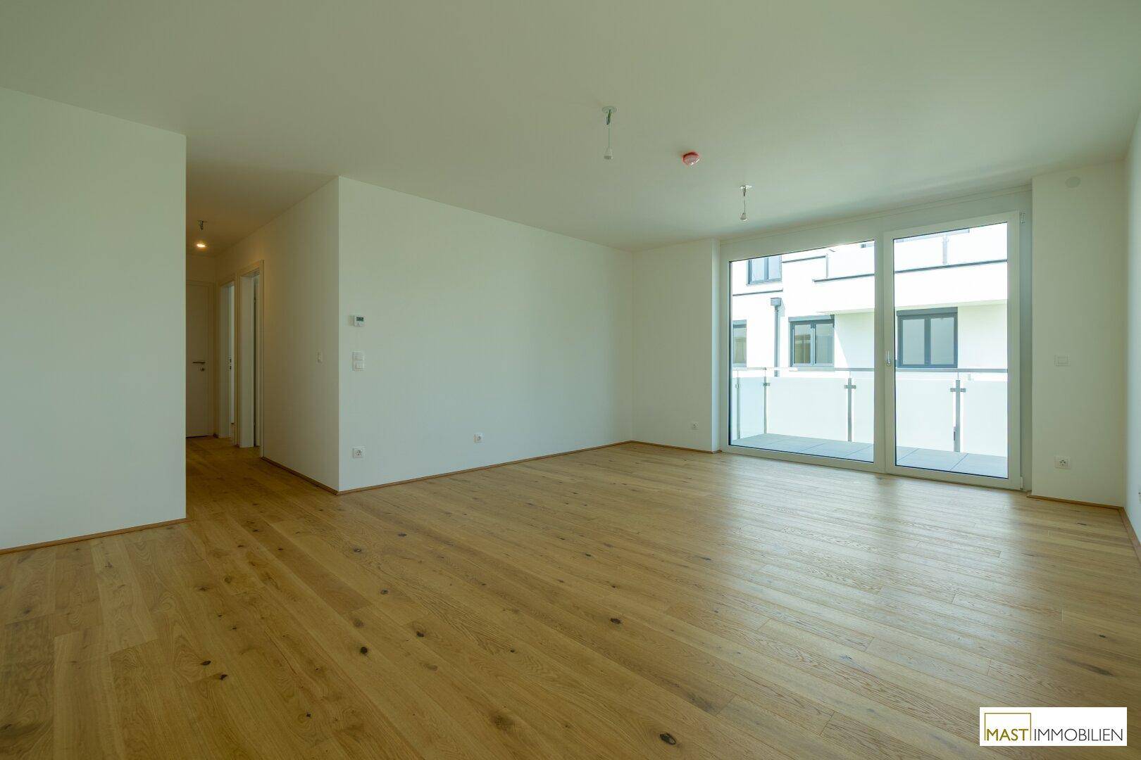 Geräumiges Wohnzimmer mit hellem Holzparkett und Zugang zum Flur sowie zum Balkon.