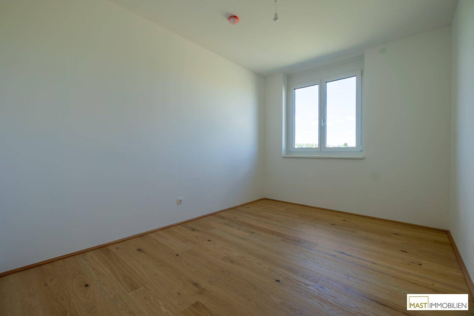 Helles Zimmer mit Holzfußboden und einem großen Fenster, ideal als Schlaf- oder Arbeitszimmer.