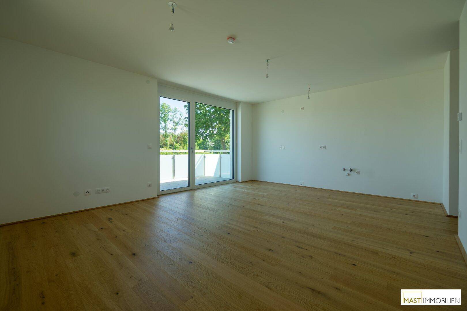 Geräumiges, leeres Wohnzimmer mit hellem Holzparkett und Zugang zu einem sonnigen Balkon.