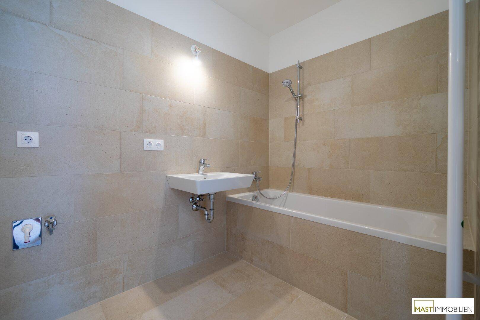 Helles Badezimmer mit beigefarbenen Wand- und Bodenfliesen, Waschbecken und Badewanne.