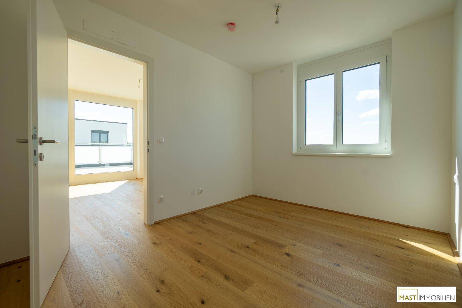 Helles Zimmer mit Holzboden und Fenster, das einen Blick auf die Terrasse bietet.