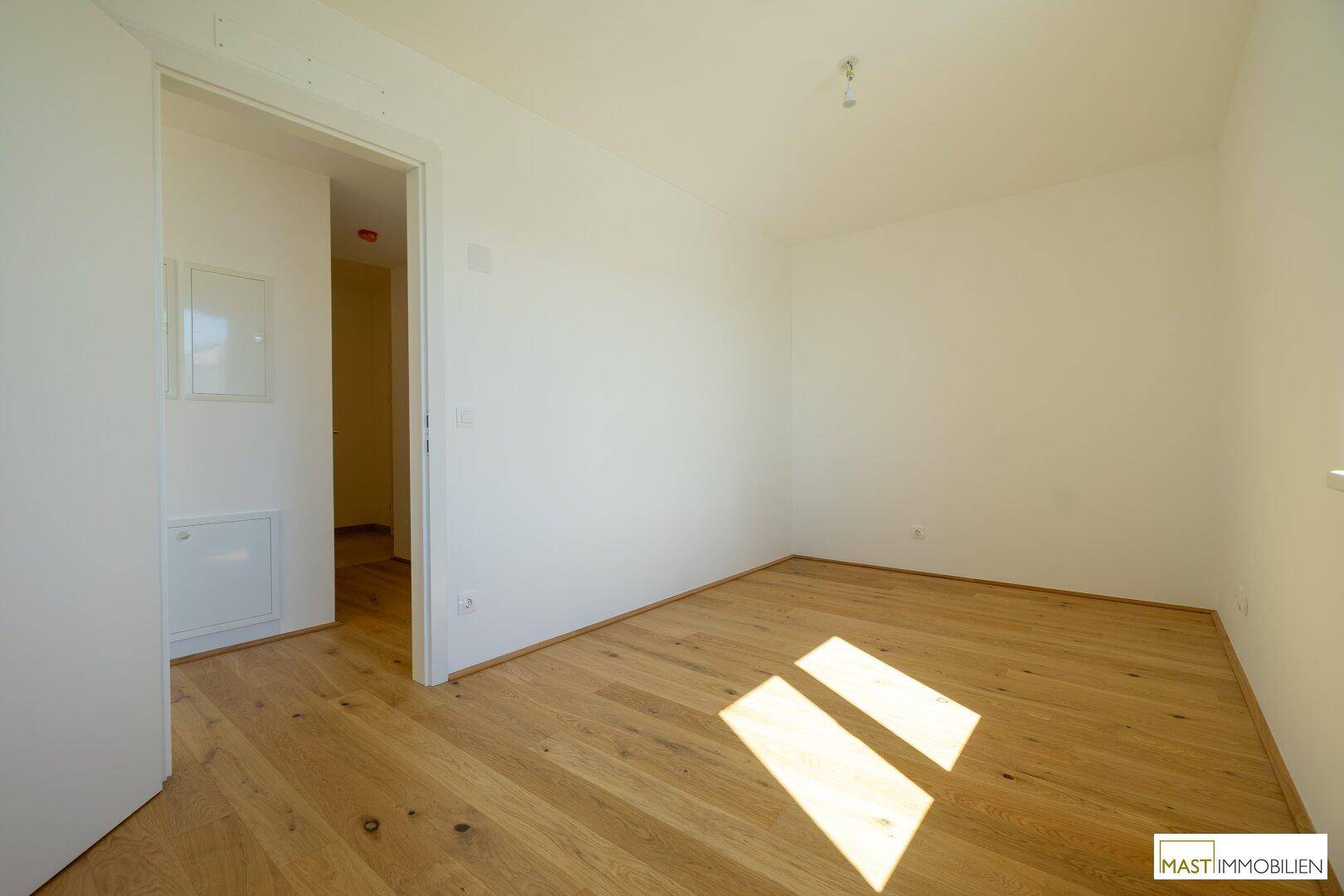 Helles, leeres Zimmer mit Holzboden und Zugang zu einem weiteren Raum.