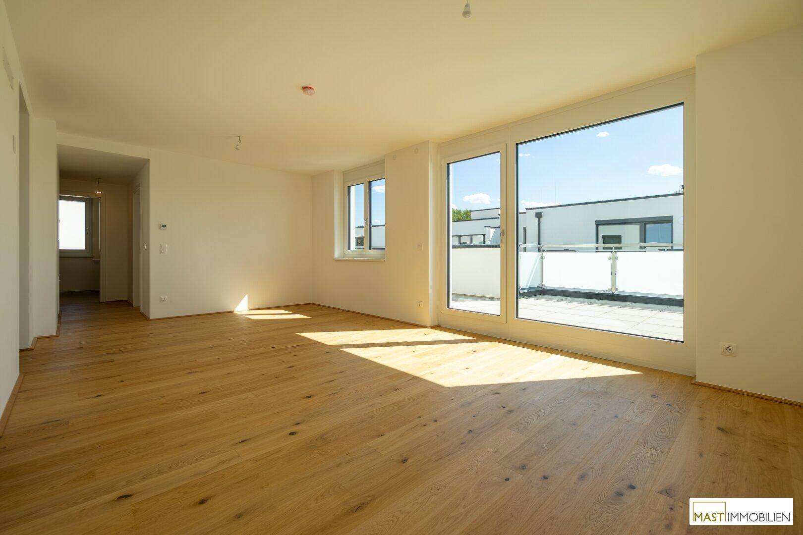 Helles, leeres Wohnzimmer mit Holzboden und großen Fenstern, die auf die Terrasse führen.