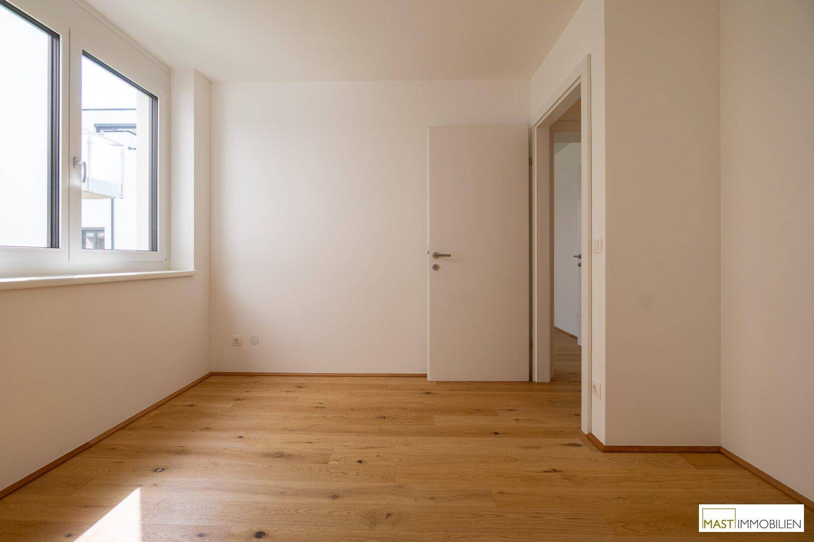 Freundliches Zimmer mit Holzboden und einem großen Fenster, das viel Tageslicht hereinlässt.