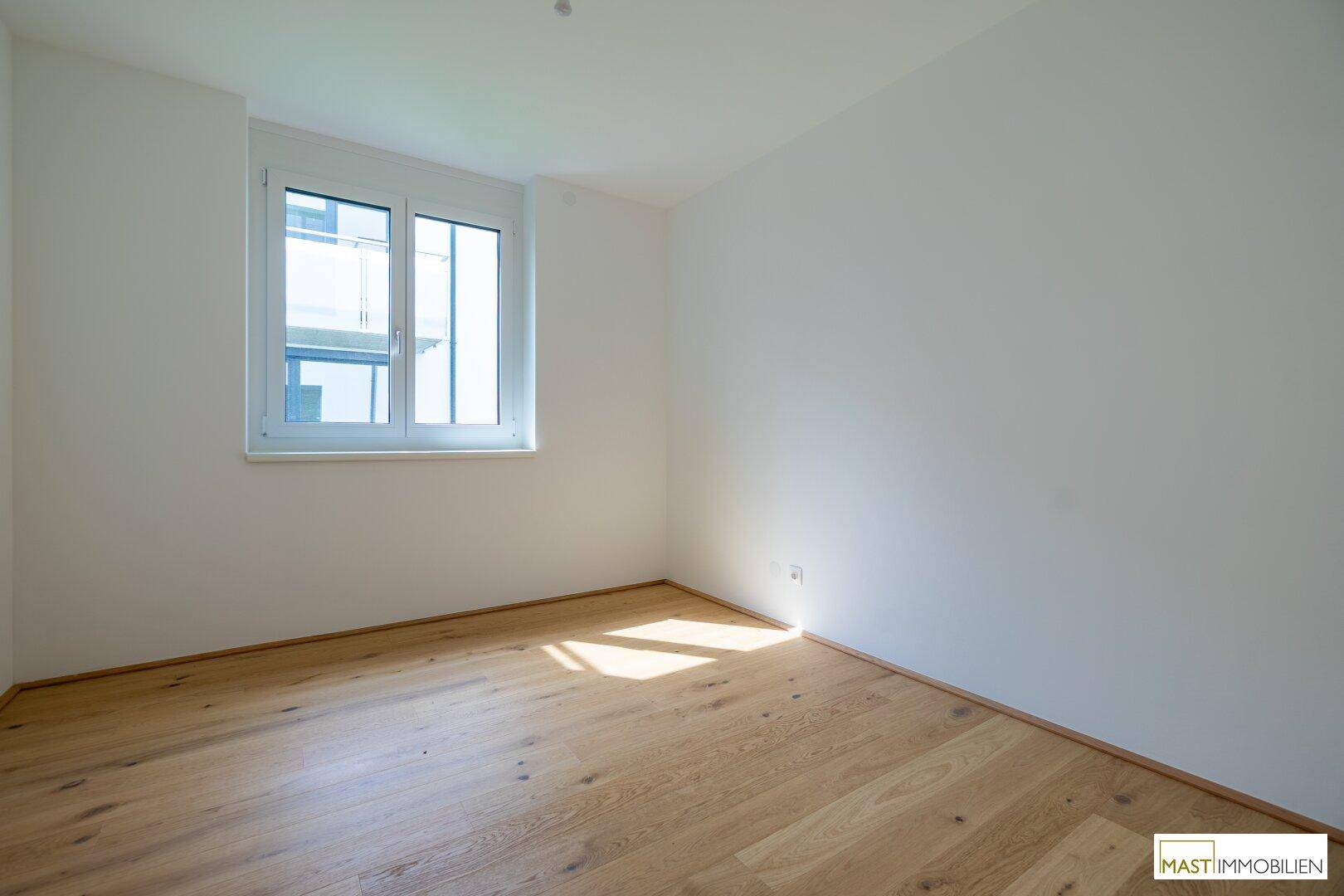 Helles Zimmer mit Holzboden und einem Fenster, das Sonnenlicht auf den Boden wirft.