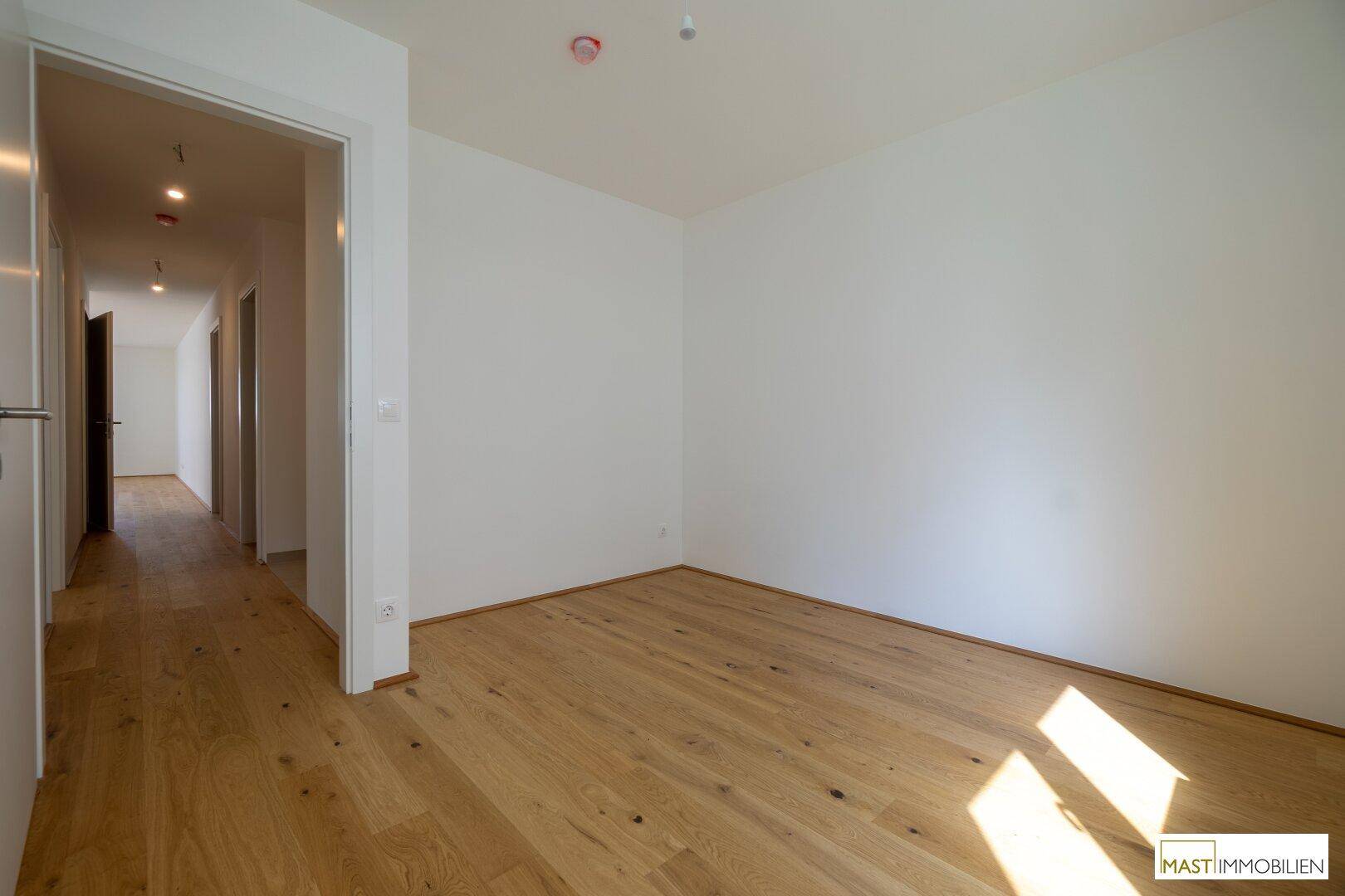 Helles Zimmer mit Holzboden und weiß gestrichenen Wänden, verbunden mit einem Flur.