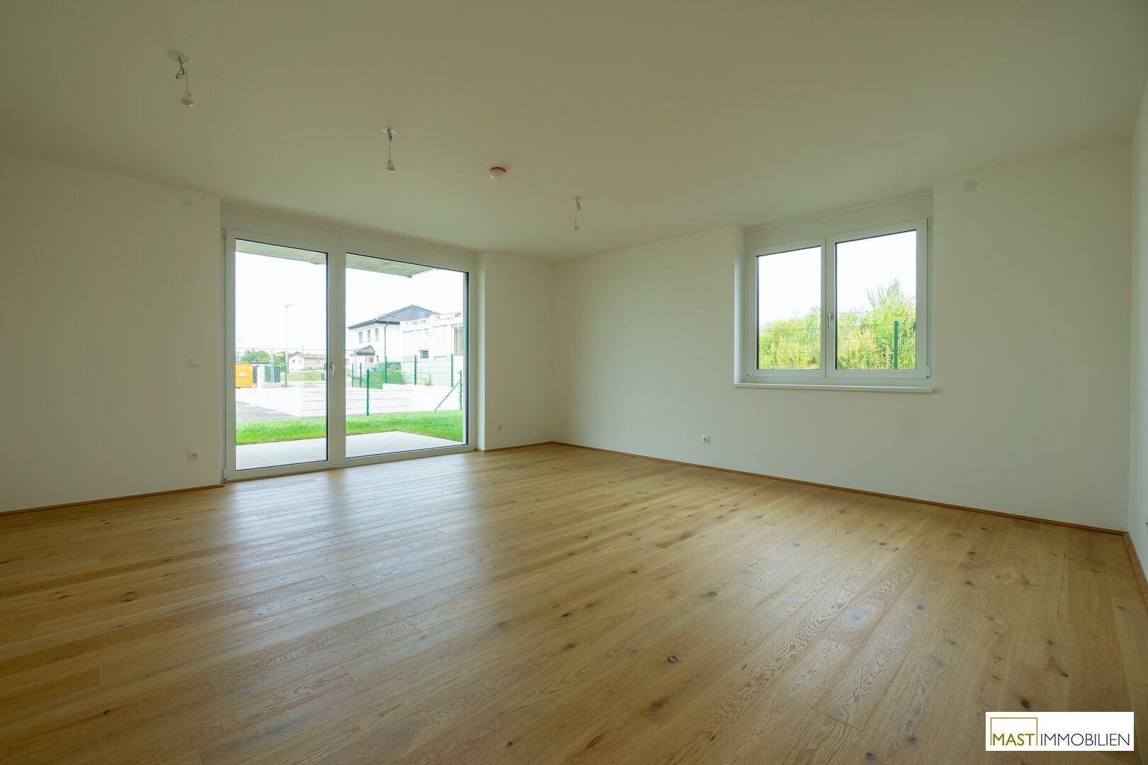 Helles, leeres Wohnzimmer mit großen Fenstern und hochwertigem Holzboden in einem Neubau.