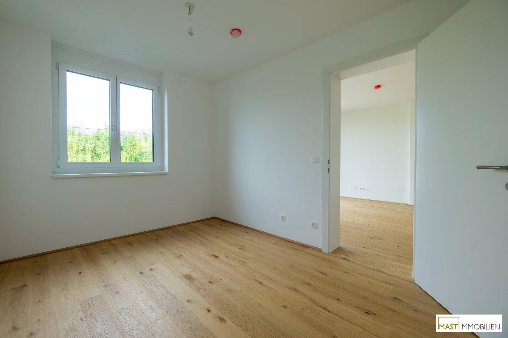 Helles Schlafzimmer mit Holzfußboden und einem Fenster, das einen Blick ins Grüne ermöglicht.