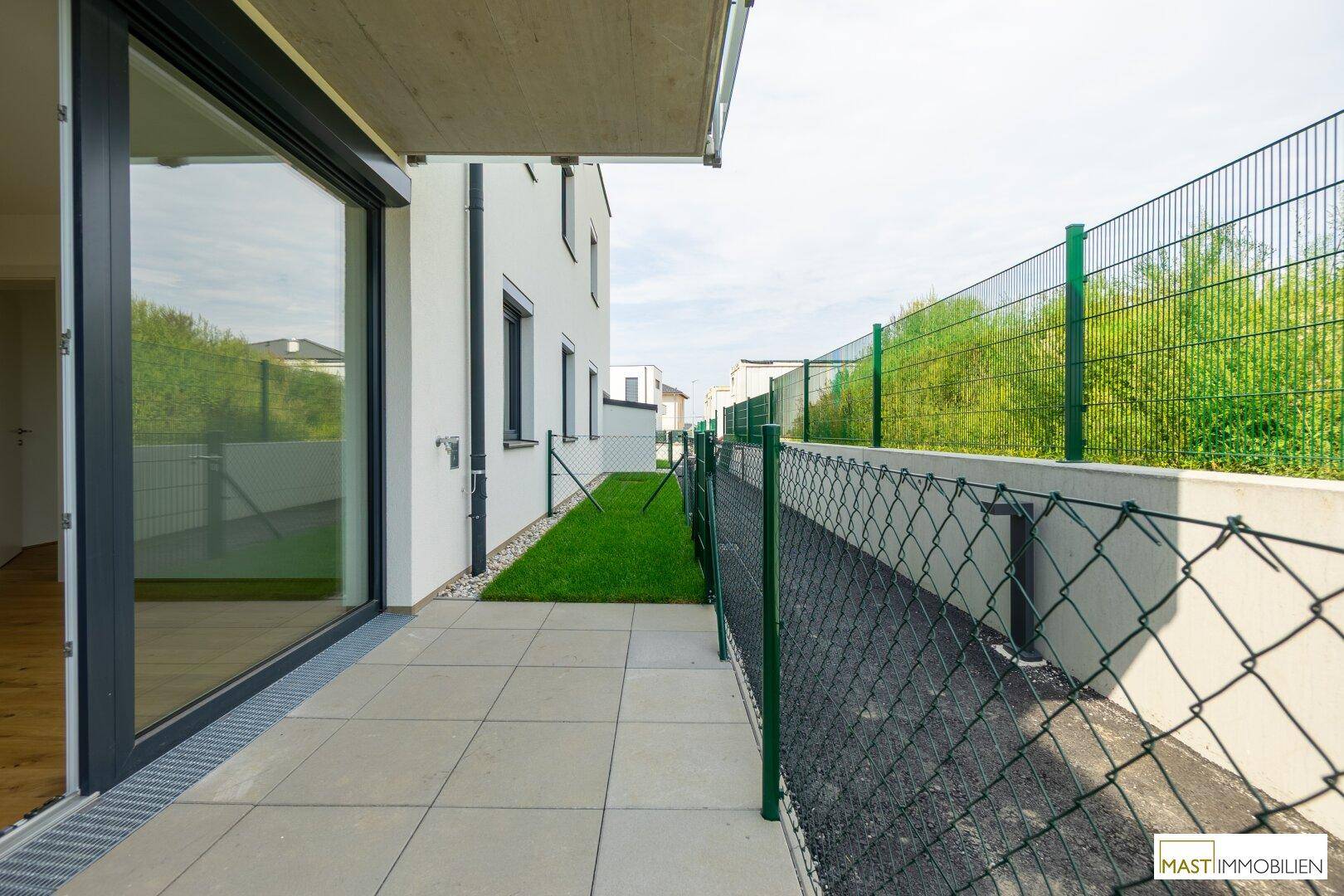 Sonniger Terrassenbereich mit Fliesenboden und kleinem Rasenstück, umgeben von einem Zaun.
