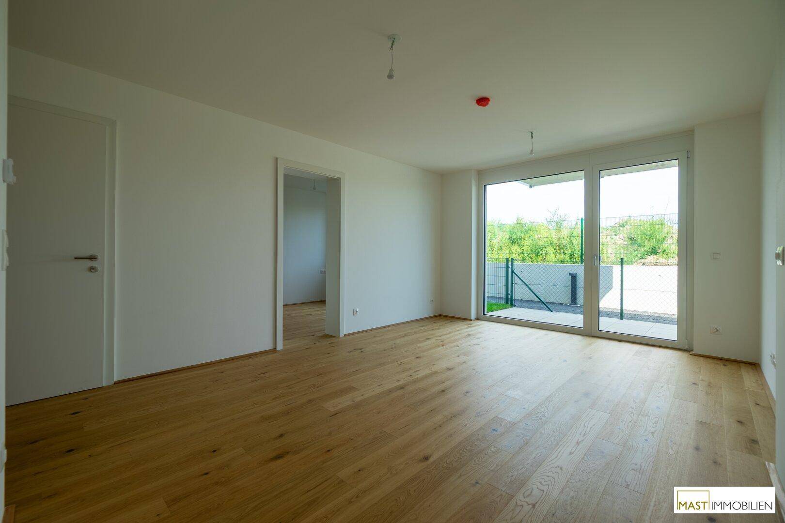 Geräumiges, helles Zimmer mit Holzboden und großen Fenstern, die Zugang zur Terrasse bieten.